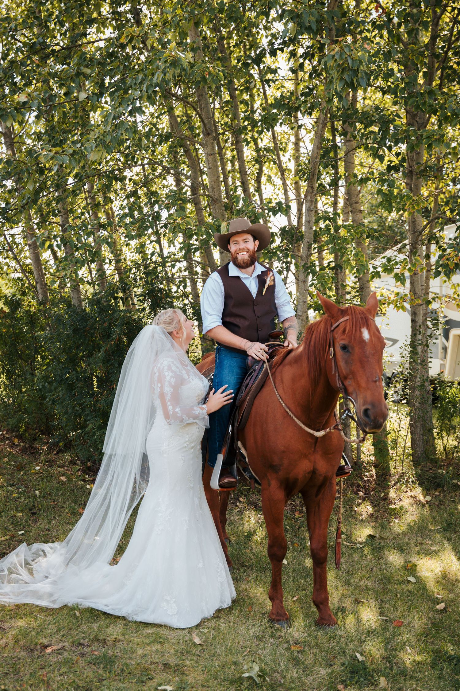 Bride in white lace gown and veil stands beside groom in cowboy hat seated on chestnut horse outdoors. Athabasca wedding photographers