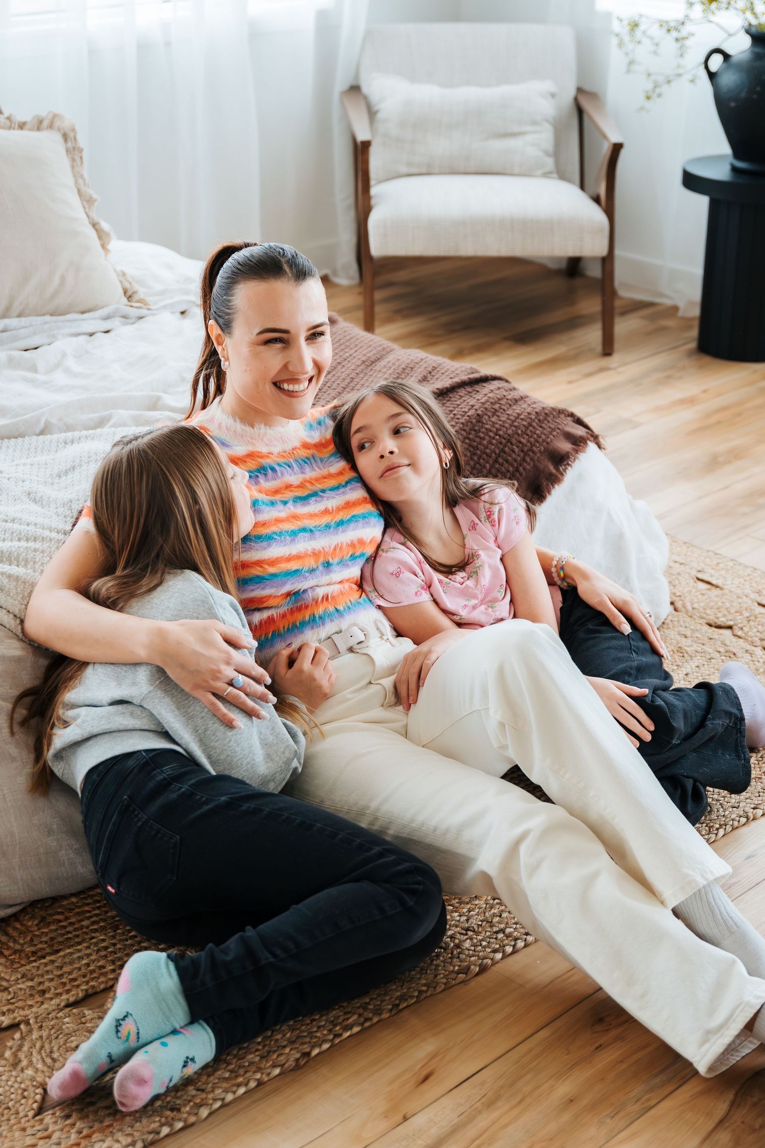 A smiling mother sitting on the floor with her two daughters leaning against a bed in a bright bedroom. Westlock Morinville Photographers