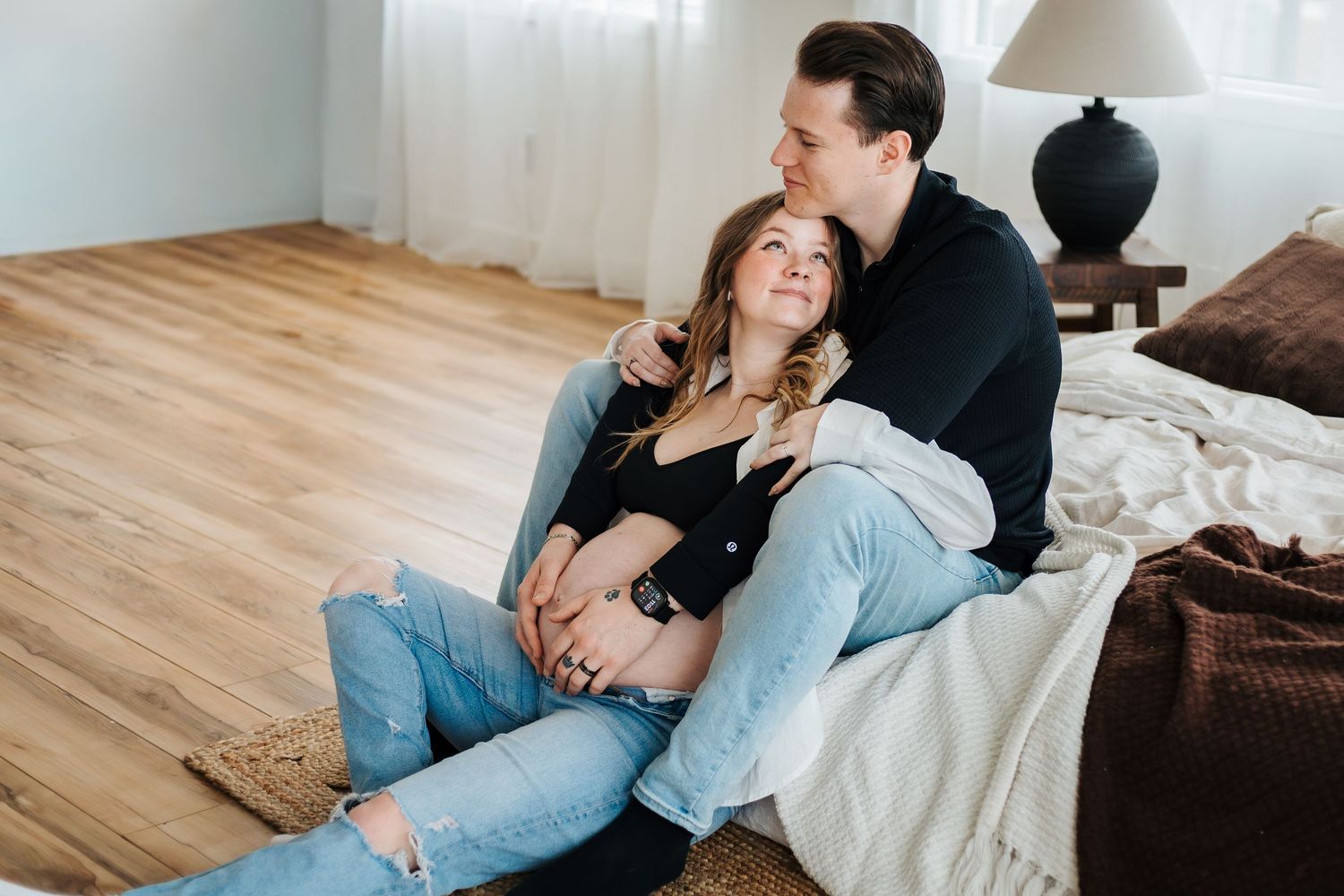 Couple sitting on floor leaning against bed, pregnant woman in jeans during a maternity photoshoot. Family and newborn photographer