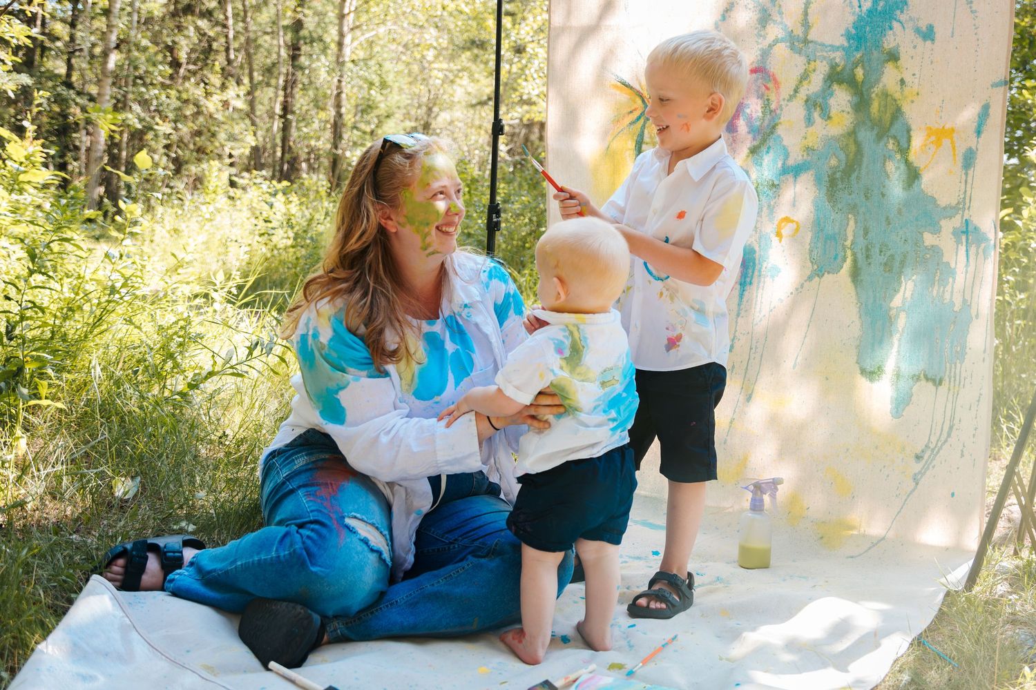 A mother sits outdoors with two young children covered in colorful paint during a fun painting session.