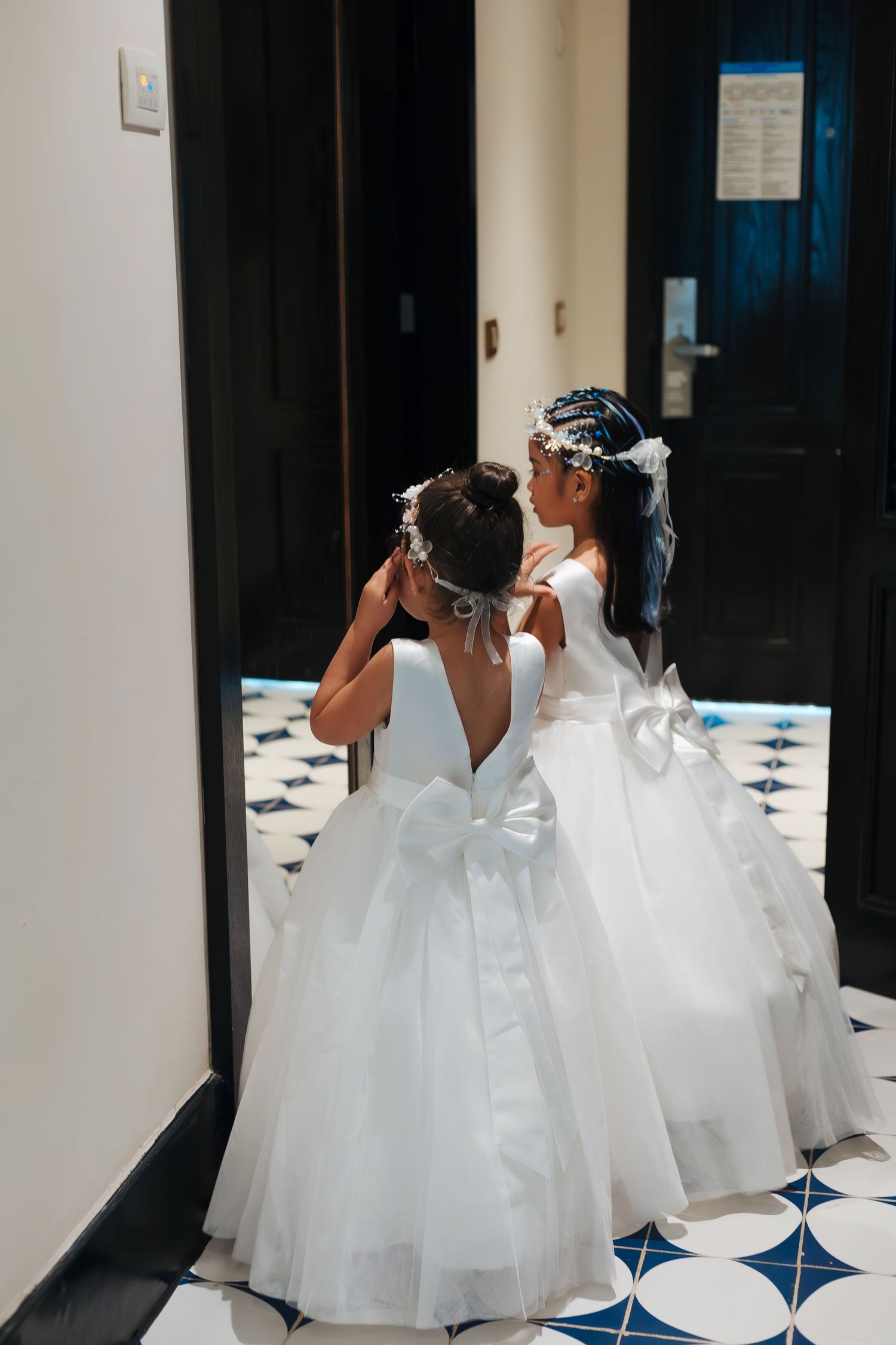 Two young flower girls in white ballgowns with bows adjust floral hair accessories in a elegant hallway.