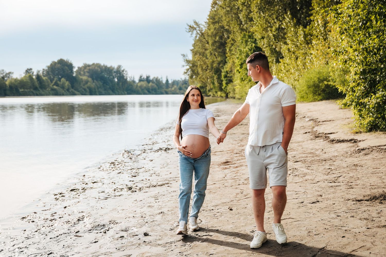 Pregnant couple holding hands walking along a sunny riverbank with lush green trees in the background. Maternity and newborn photographer