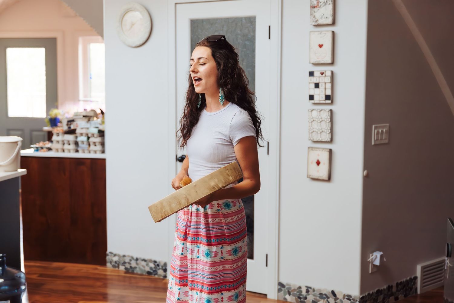 Woman laughing while holding a rolled yoga mat in a bright home interior, wearing a white top and colorful skirt.