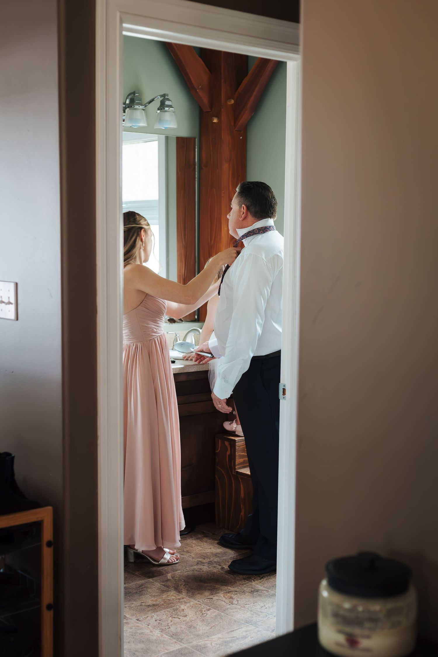 A woman in a pink gown helps a man in a white shirt tie his tie in a rustic bathroom before a wedding.