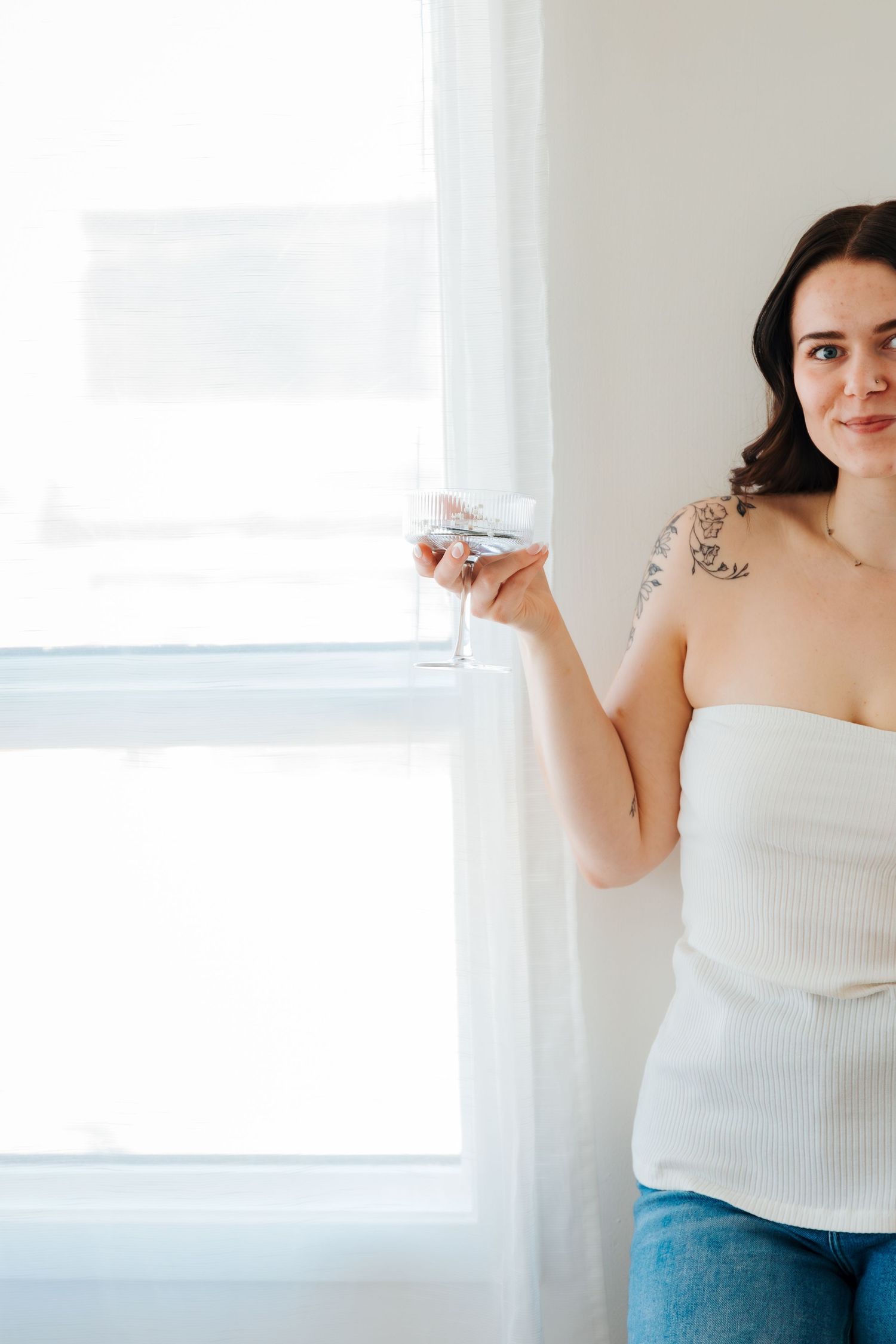 Woman in white tube top with floral shoulder tattoo holding a glass near a bright window. St.albert business photography.