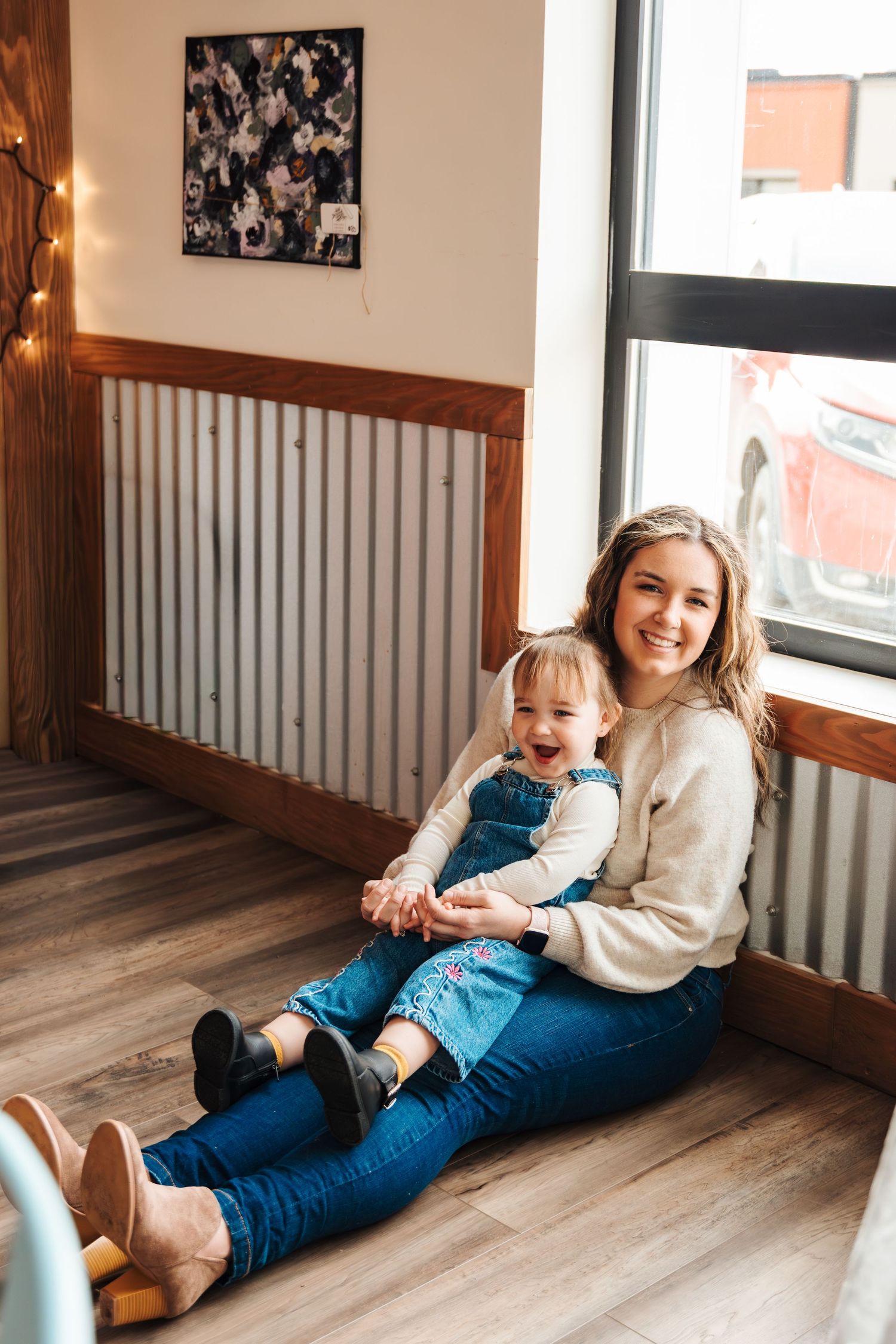 Young woman sitting on floor holding smiling toddler girl in denim dress near window in cozy room. photography studio calgary