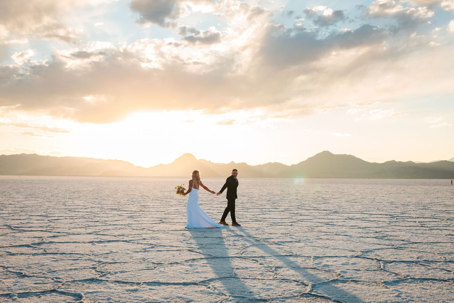 Bride and groom hold hands at sunset on the Bonneville Salt Flats, with mountains and golden sky behind them. Destination wedding mexico
