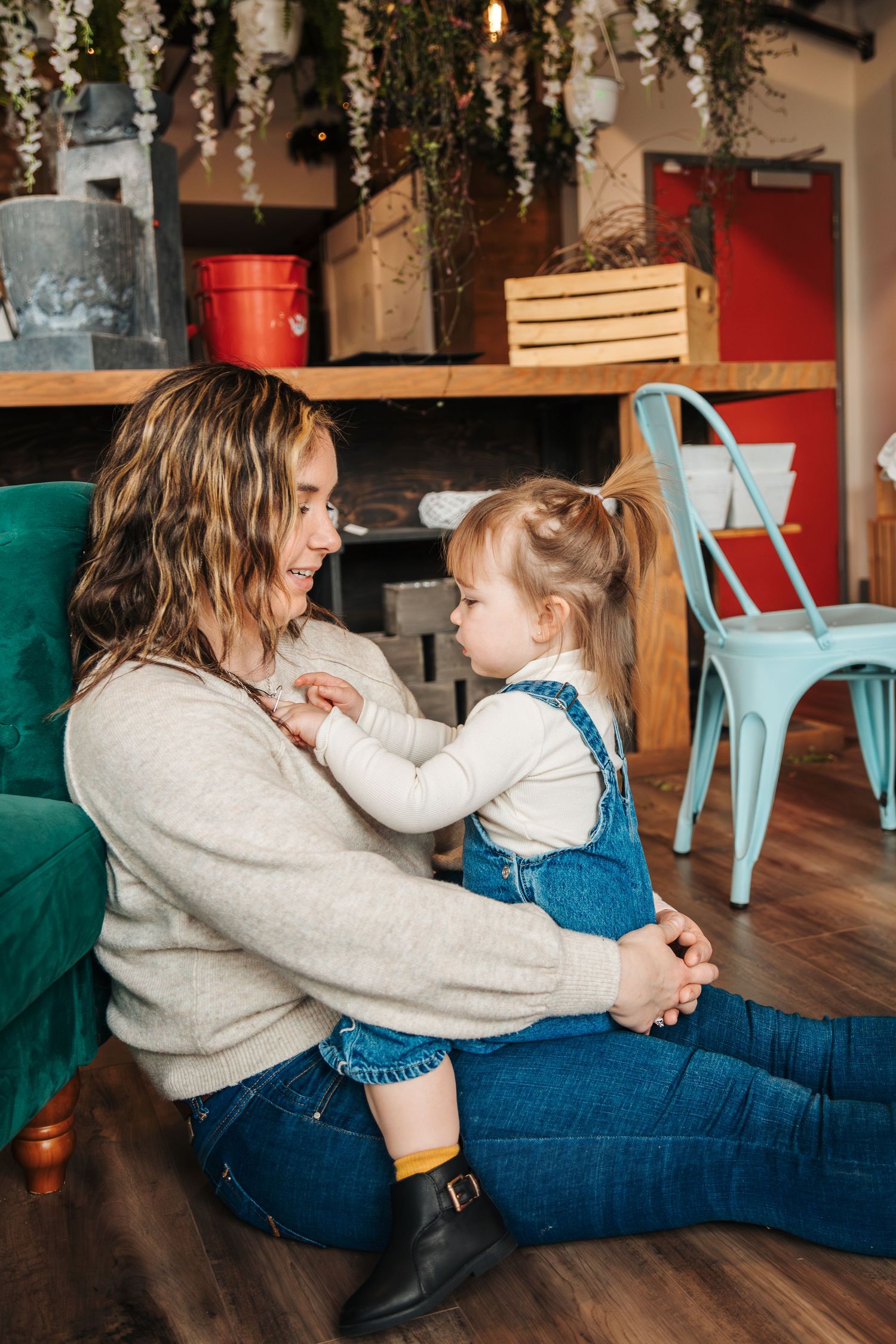 Mother and toddler girl sitting on floor, smiling and playing together in a cozy cafe with teal chairs. Lady Bugs Flowers coffee shop
