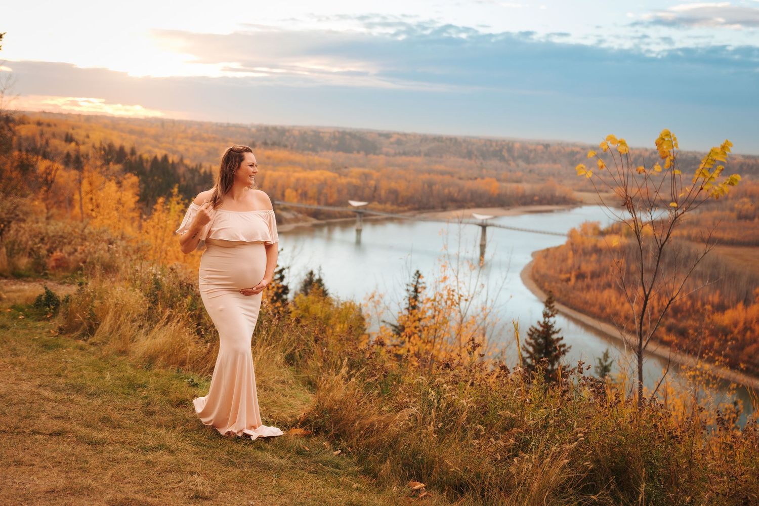 Pregnant woman in pink gown stands on a hillside in Edmonton overlooking a winding river at golden hour. Maternity photo session
