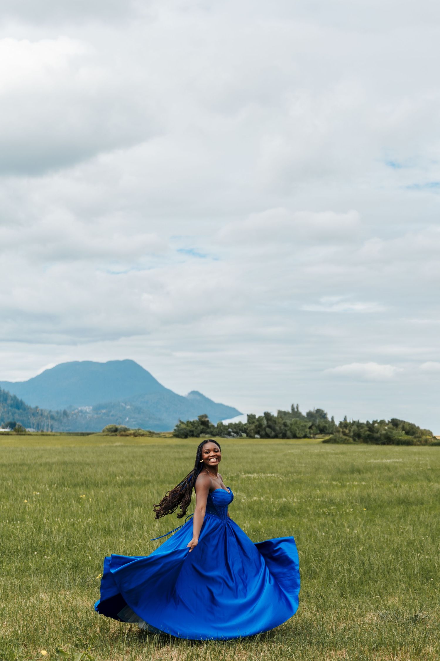 A joyful woman in a flowing royal blue gown twirls in a green field with mountains in the background. Graduation pictures. family pictures.