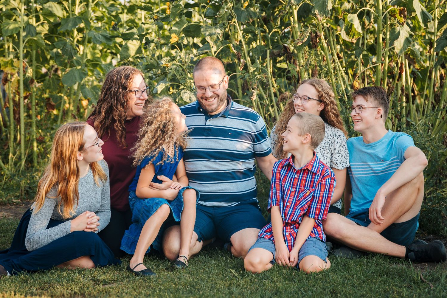 A family of seven sits together laughing on green grass in front of sunflowers on a sunny day.