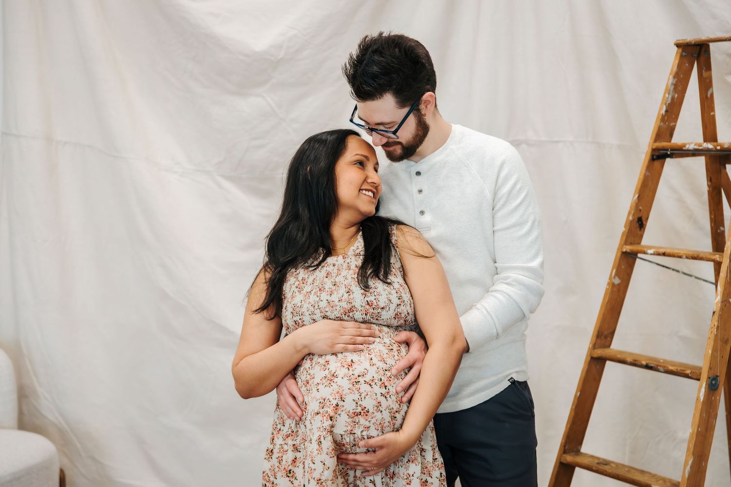 Couple sharing a tender maternity photo shoot moment, man embracing pregnant woman in floral dress against white draped backdrop. family
