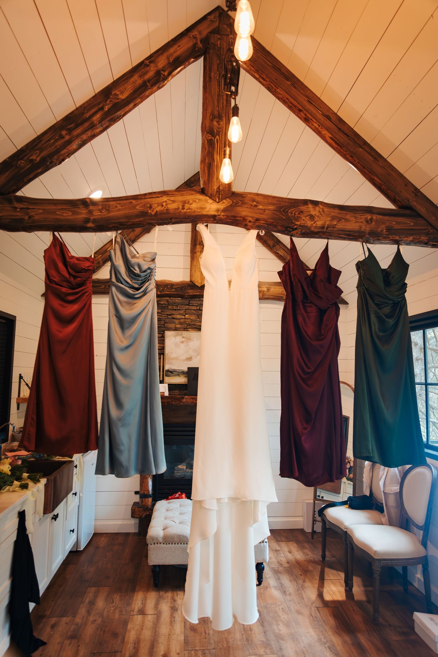 Wedding dress and colorful bridesmaid gowns hanging from rustic wood beam ceiling in a cozy bridal suite. The valley weddings venue alberta
