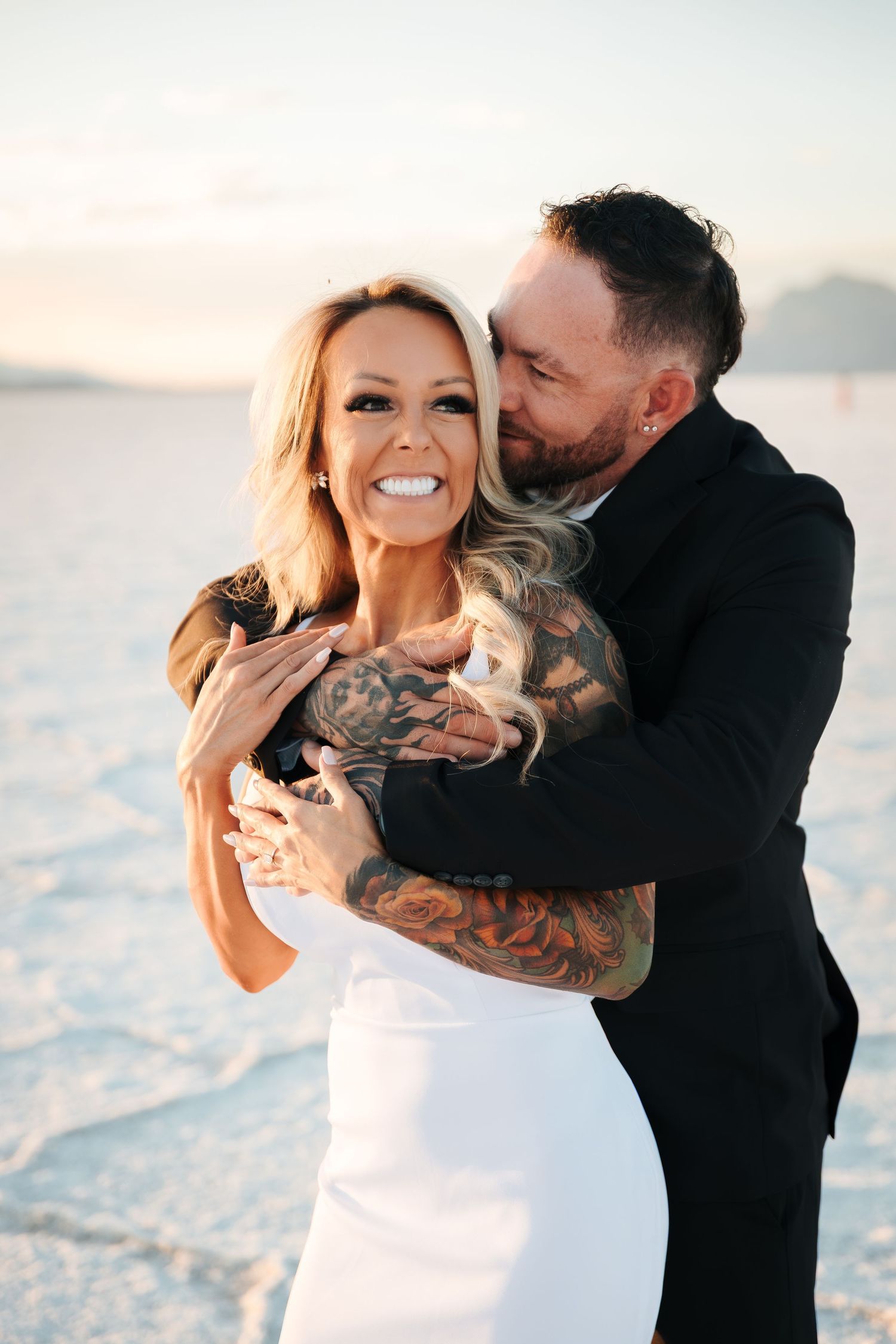 Couple embracing at sunset on white salt flats, bride in white dress smiling, groom in black suit with tattoos. golden hour wedding photos