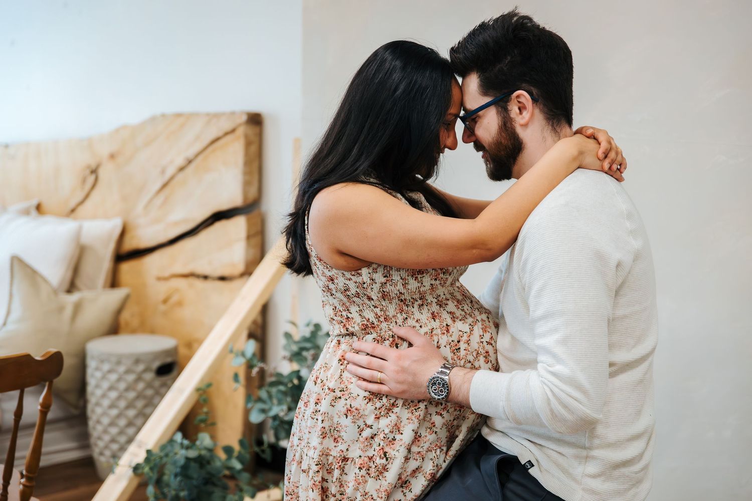 A couple embraces tenderly in a cozy bedroom, the woman in a floral dress with her arms around the mans neck. stillbirth photos