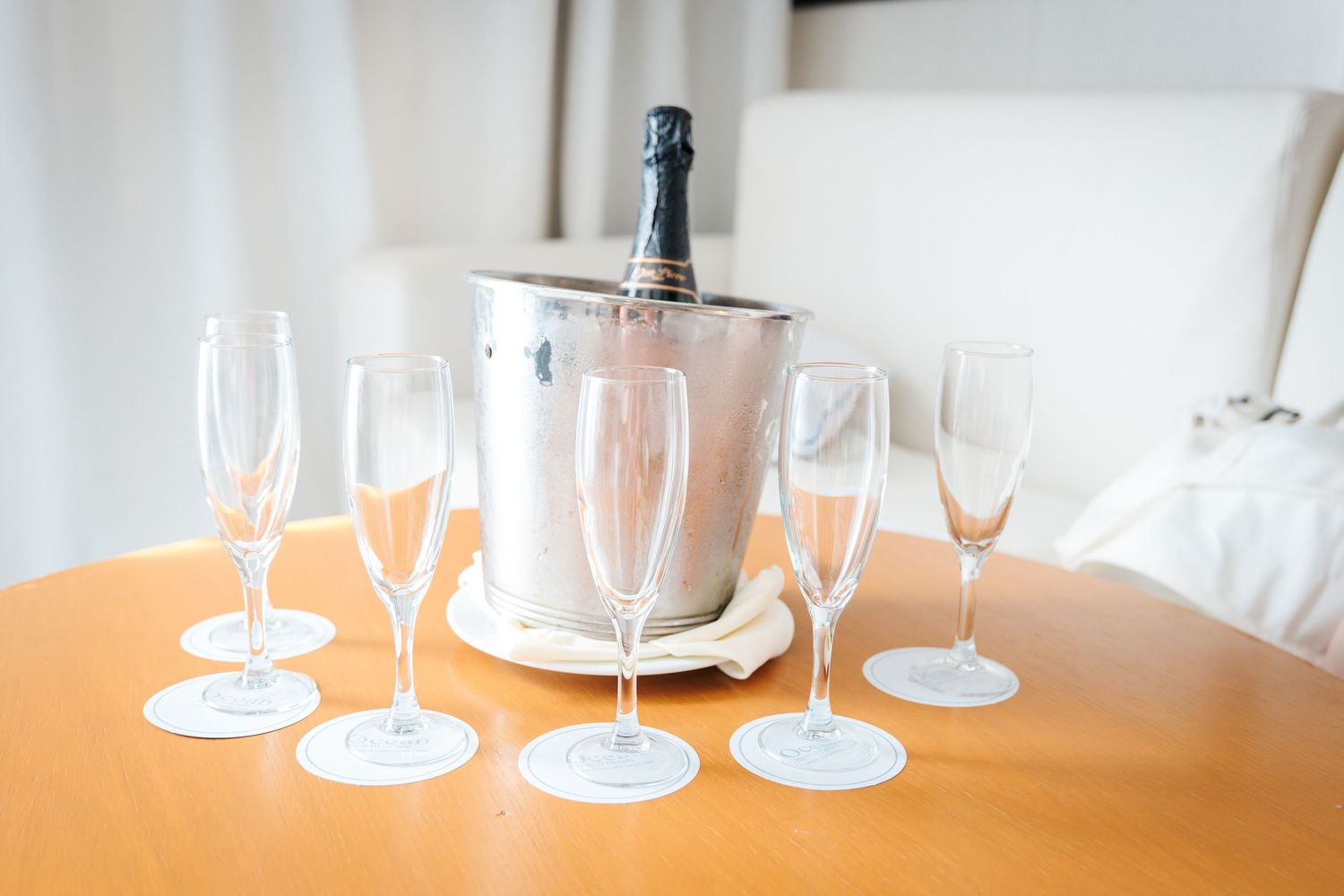 Five empty champagne flutes arranged around a silver ice bucket with a champagne bottle on a wooden table.