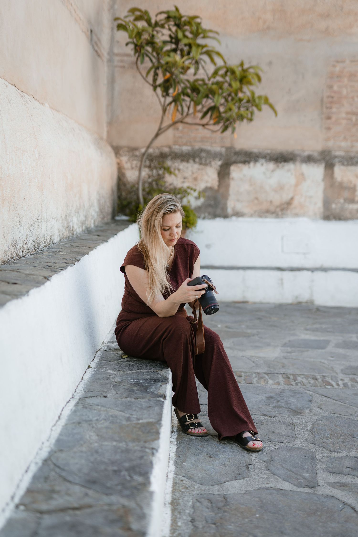 Blonde woman in brown jumpsuit sitting on stone steps reviewing photos on a DSLR camera outdoors.