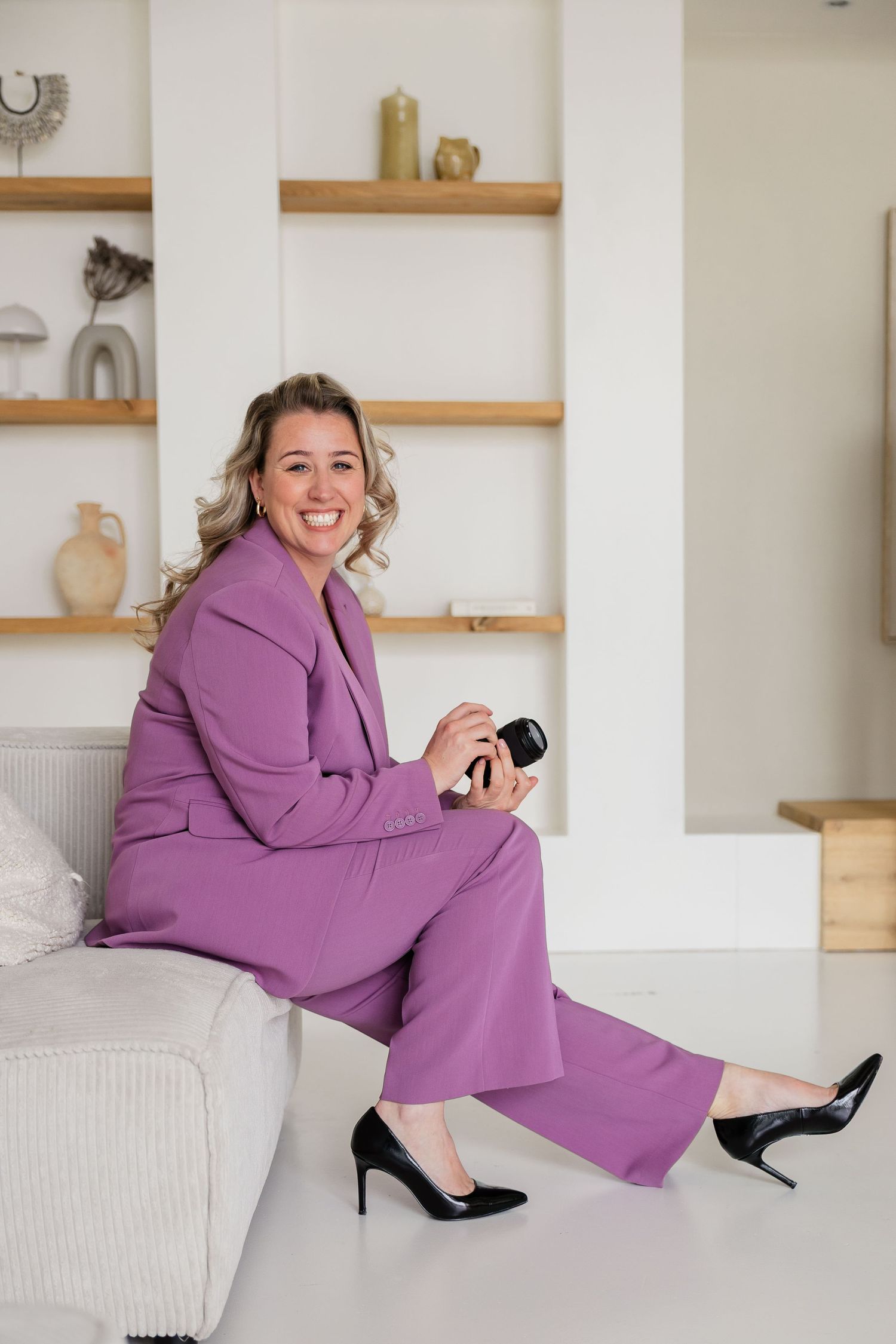 Woman in purple suit sitting on sofa holding camera, smiling in bright white interior with wooden shelves.