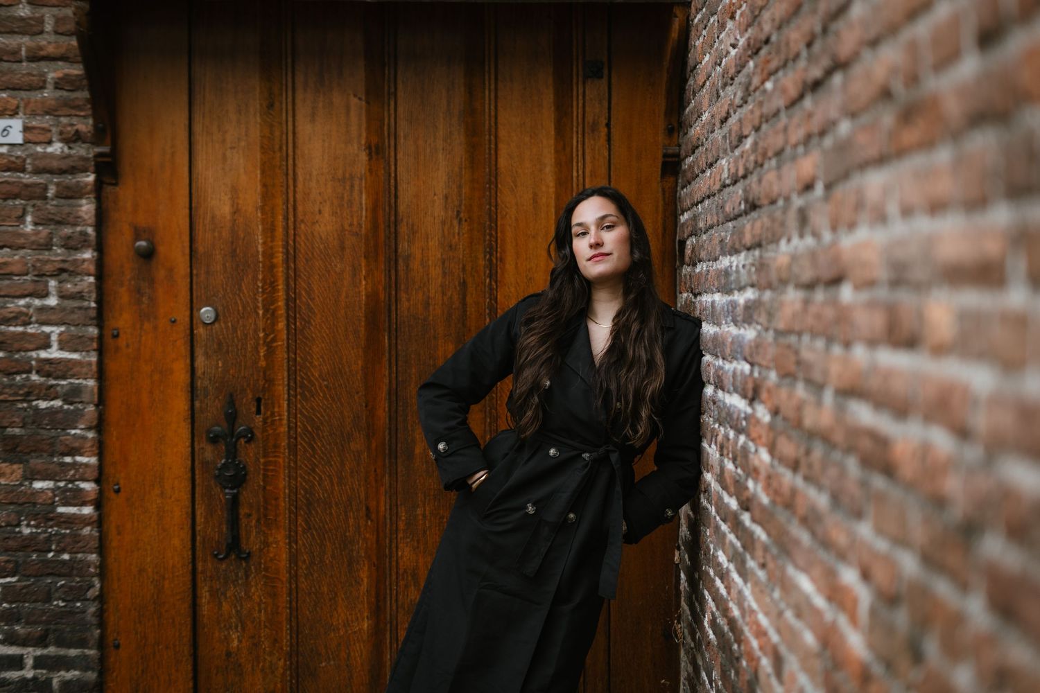 Woman in black coat leaning against brick wall near wooden door.