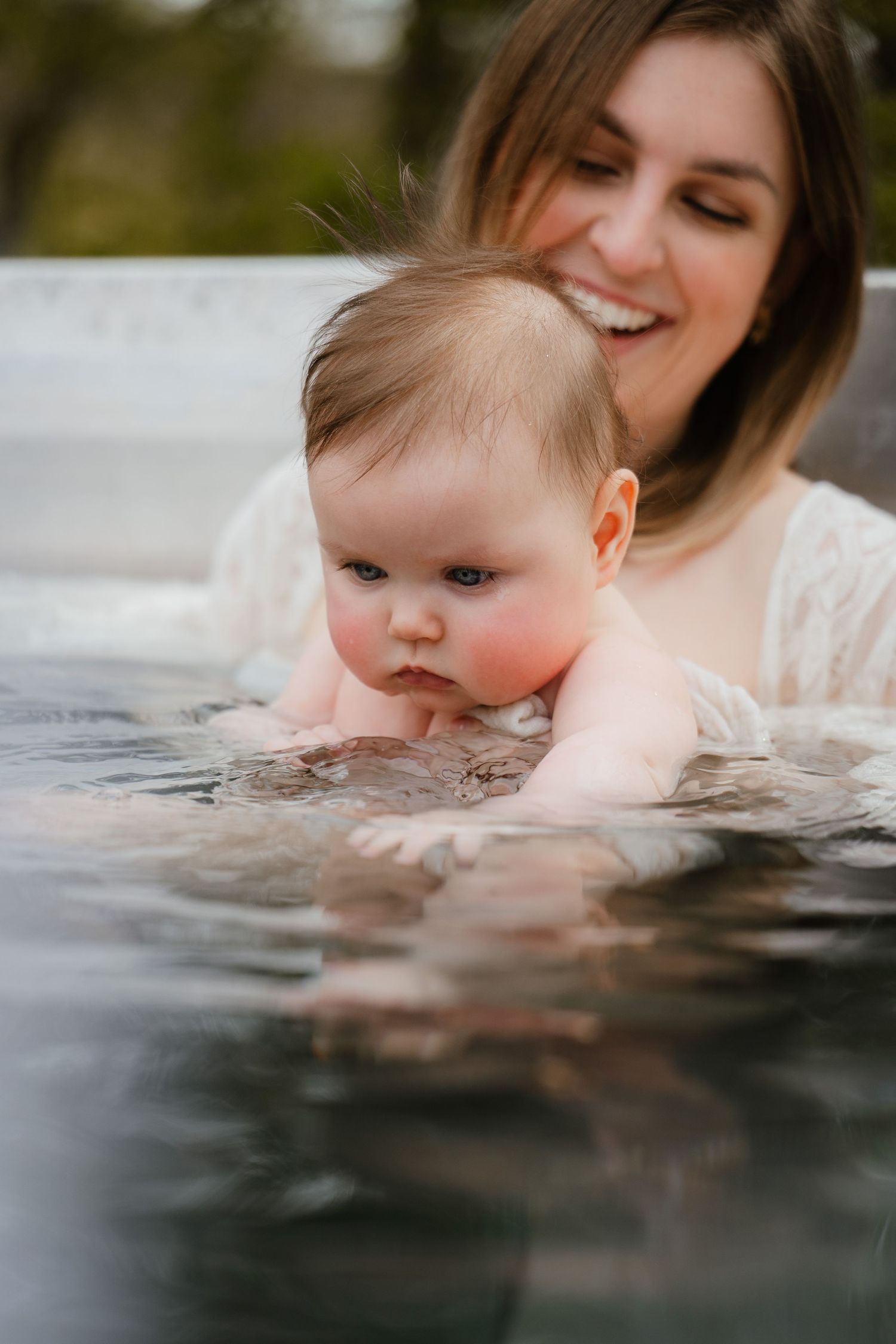 A smiling mother holds her baby in a pool, introducing the infant to water in a tender moment.