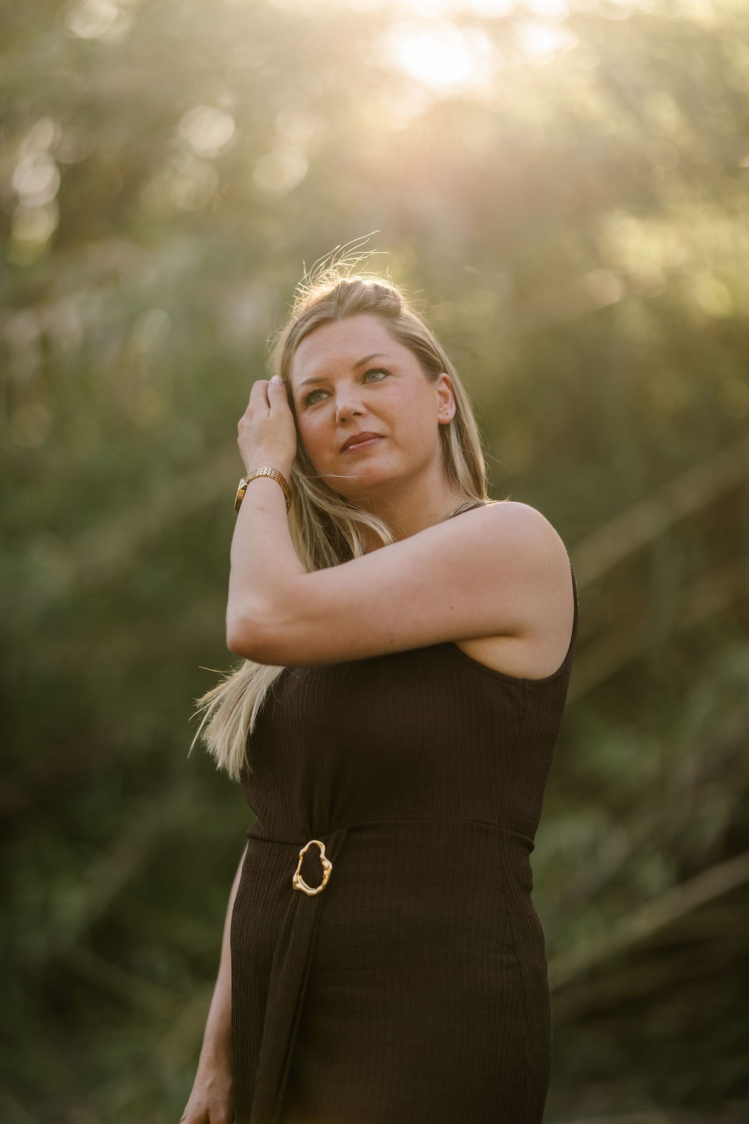 Blonde woman in black sleeveless dress posing in sunlit forest with warm golden light.
