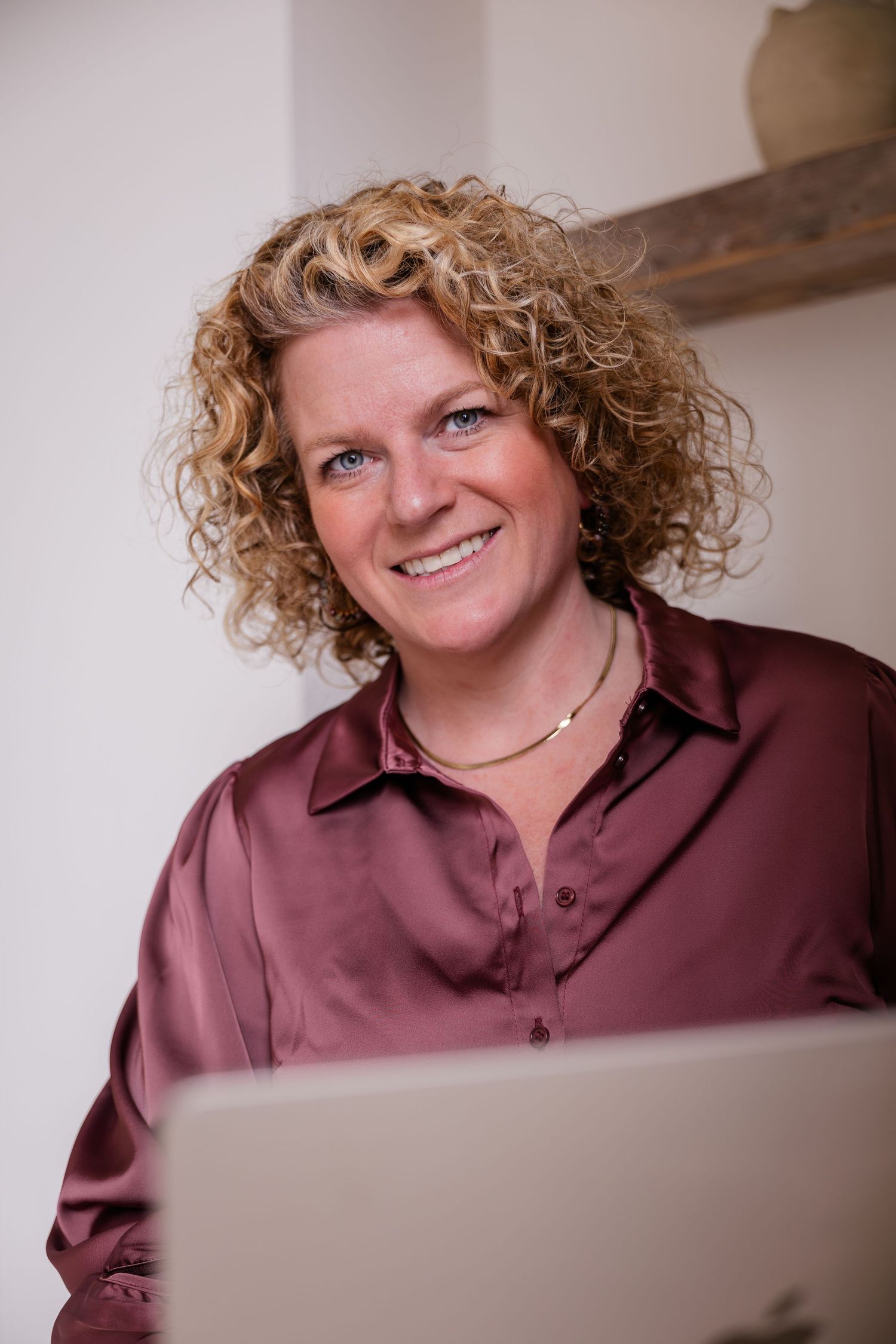 Smiling curly-haired woman in burgundy silk shirt working on laptop in bright interior.