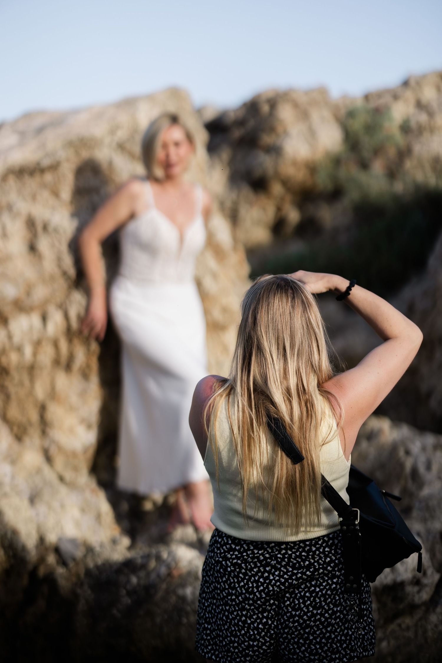 Photographer taking pictures of blonde woman in white dress posing against rocky cliff backdrop.