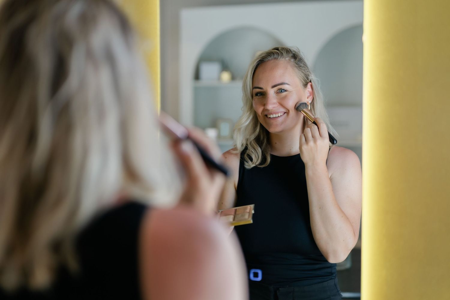 Lachende blonde vrouw die make-up aanbrengt met een kwast terwijl ze in een spiegel met gele lijst kijkt.