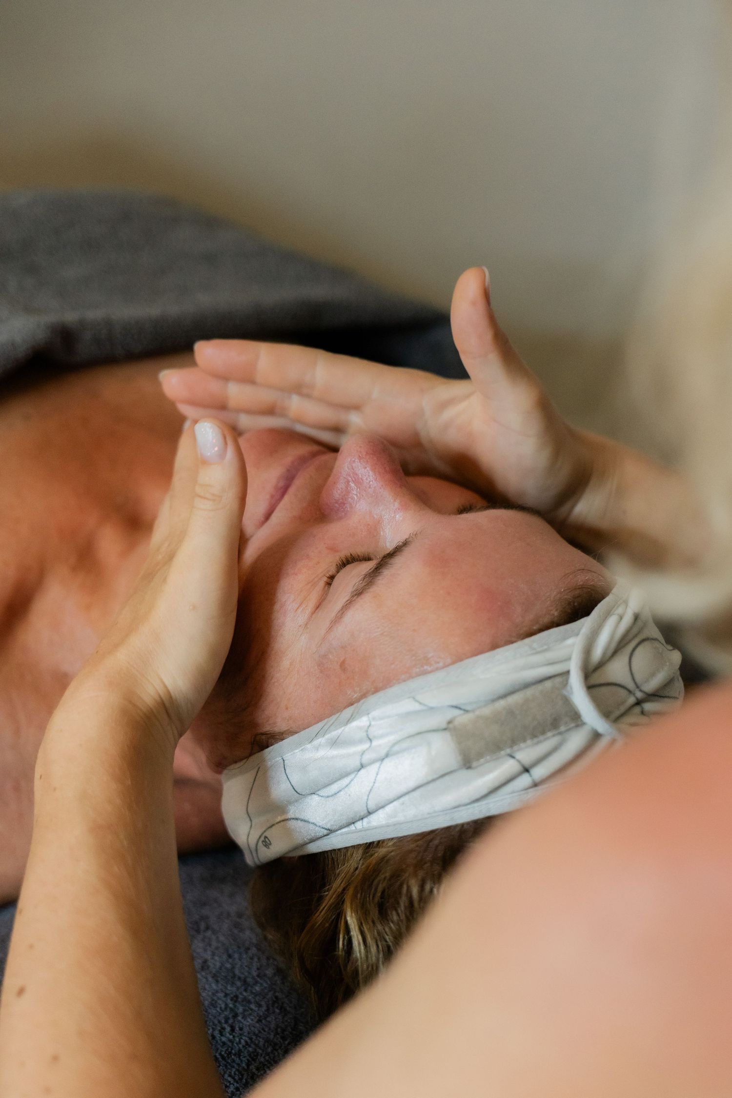 Esthetician performing a gentle facial massage on a relaxed woman wearing a headband on a table.