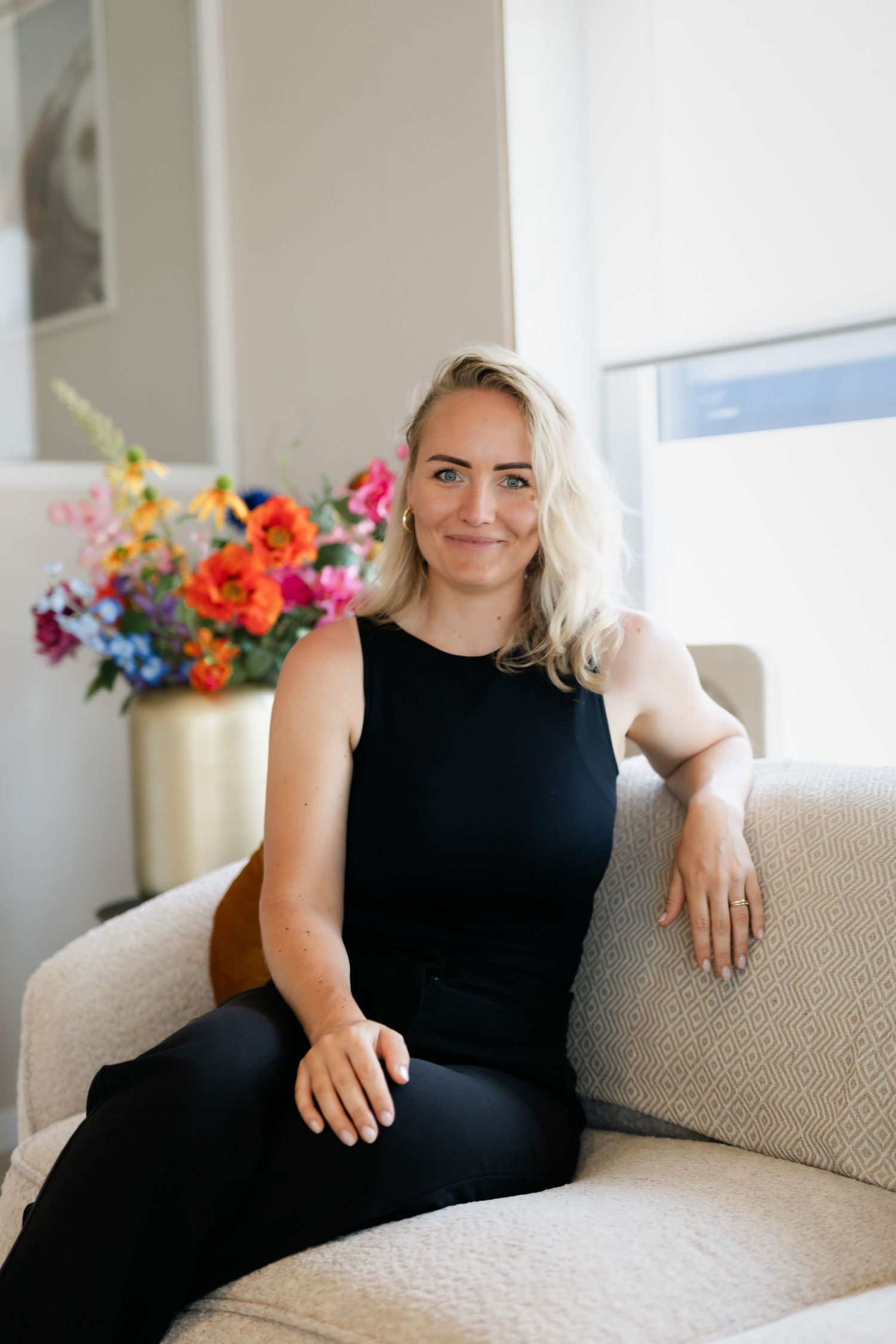 Smiling blonde woman in black outfit relaxing on cream sofa with vibrant floral arrangement.