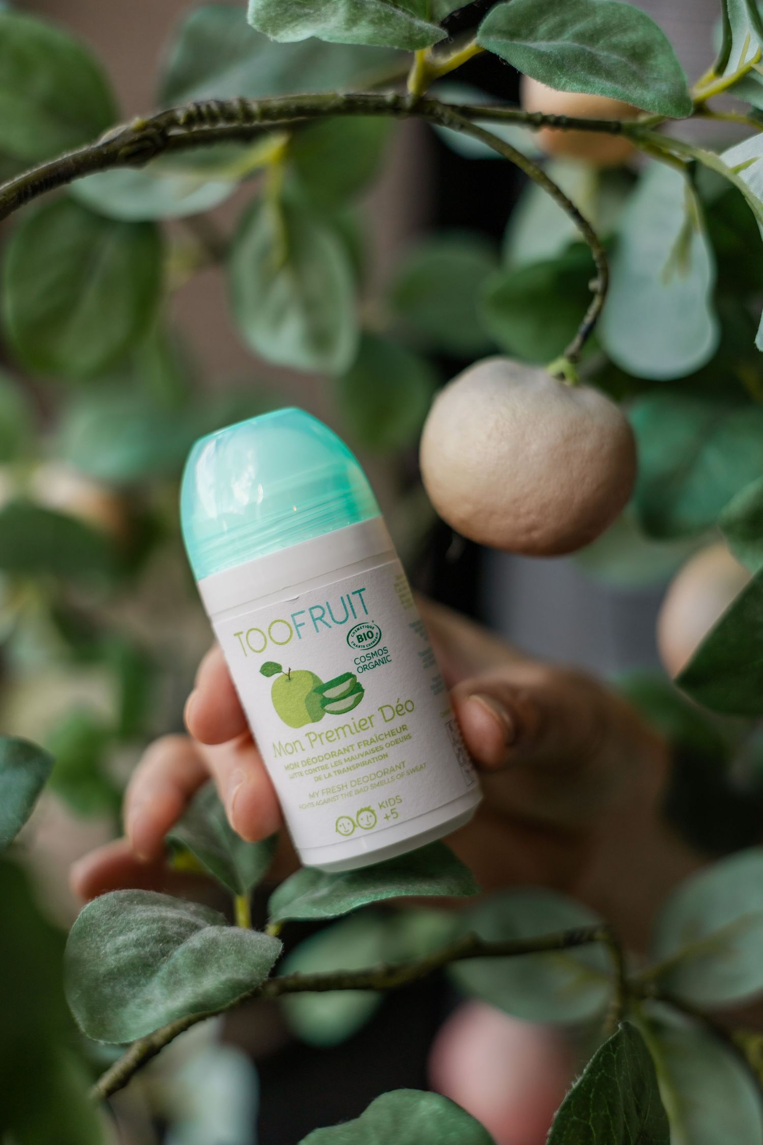 Hand holding TOOFRUIT Mon Premier Déo organic roll-on deodorant in a leafy green tree with fruit.