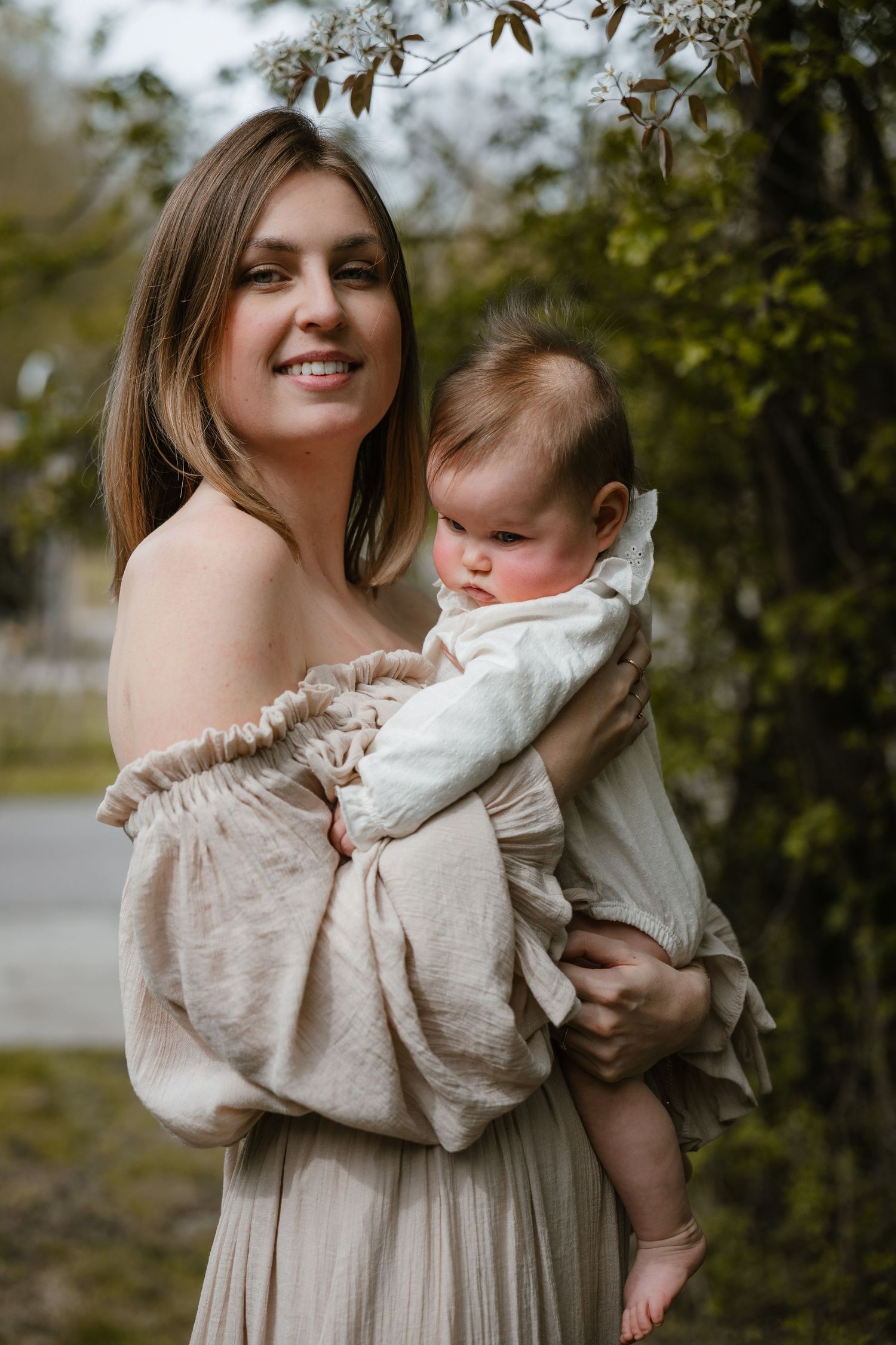 Smiling mother in beige off-shoulder dress holding infant baby outdoors near blooming trees.