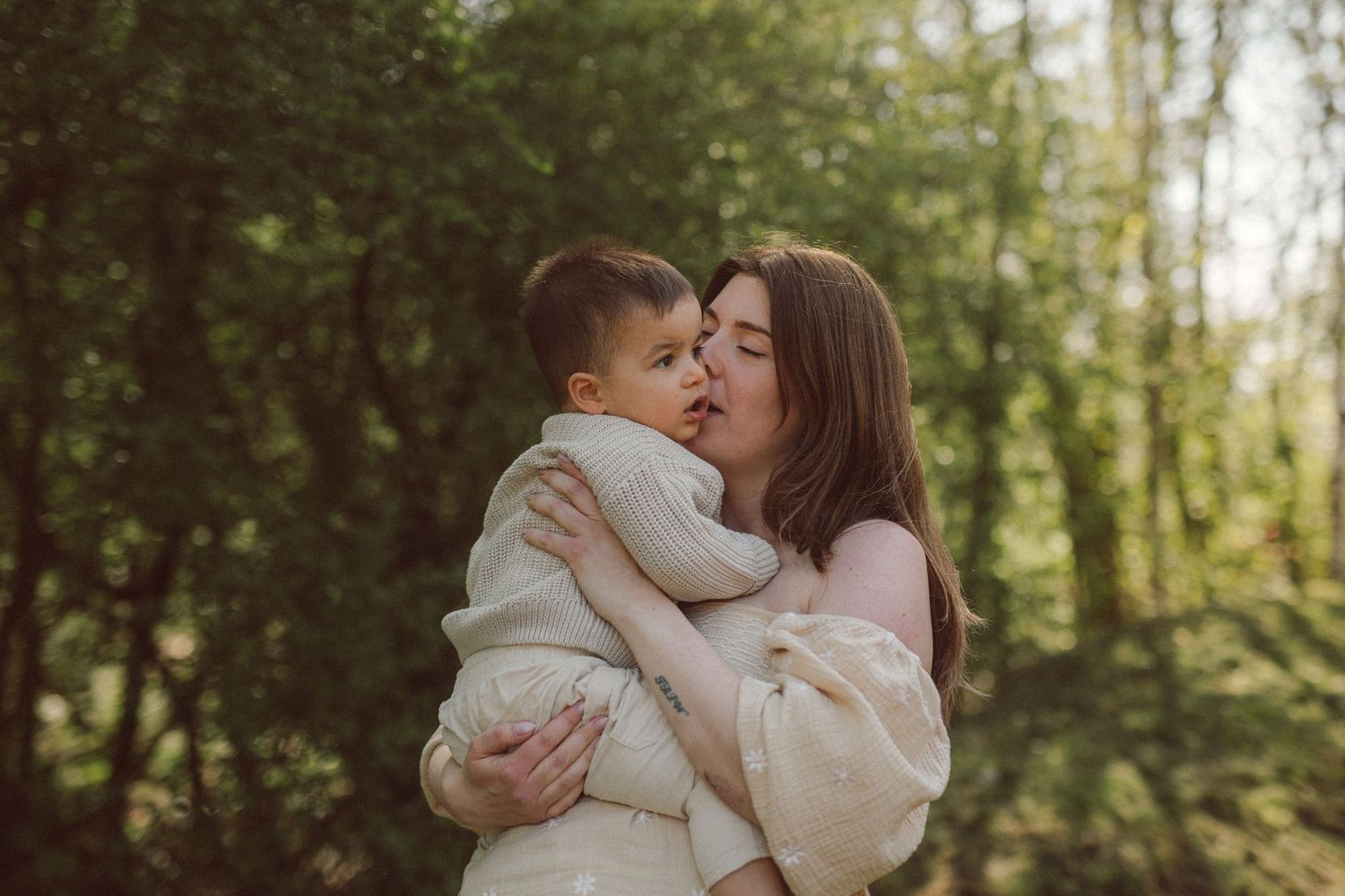 Loving mother hugging and kissing young child in lush green forest.