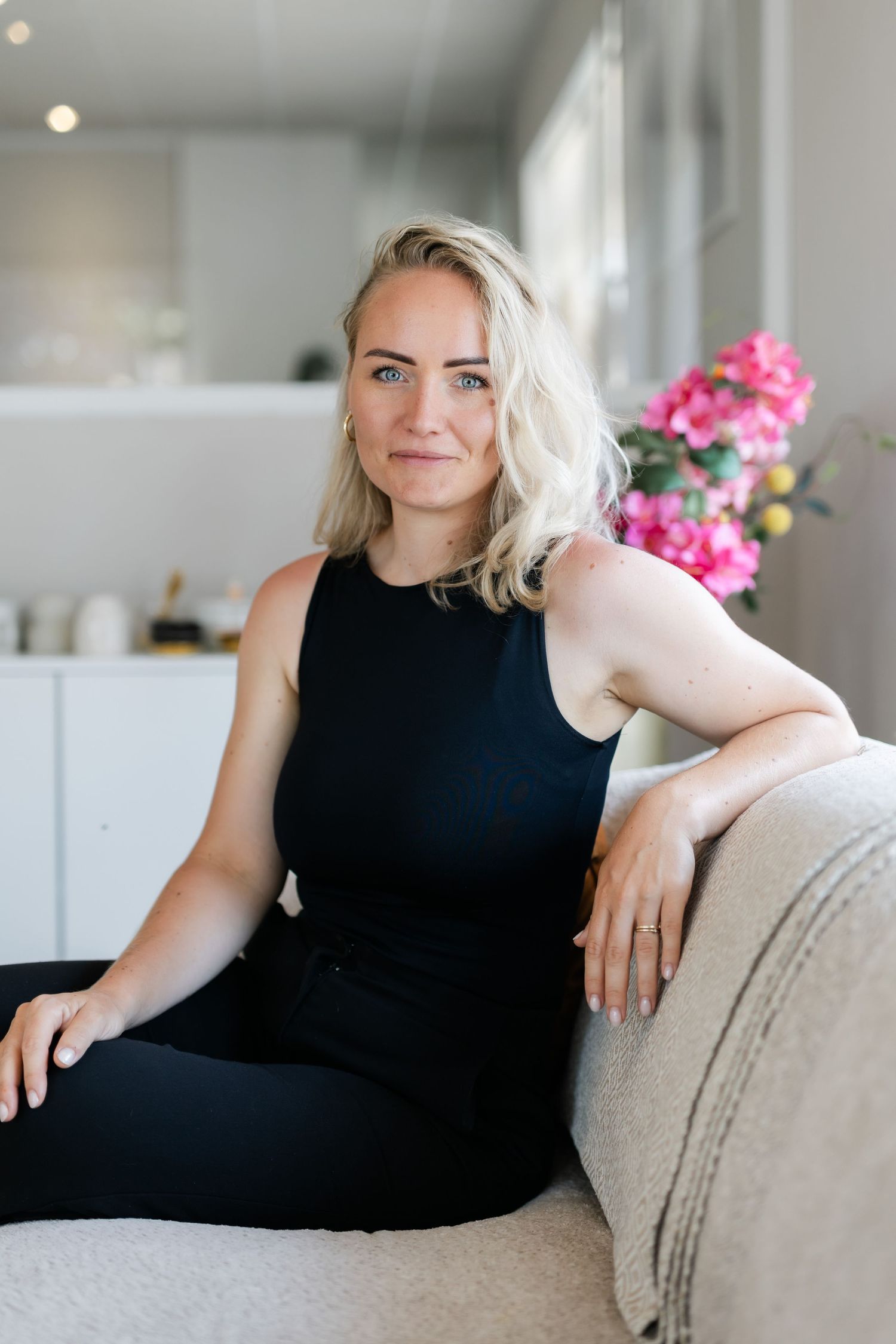 Smiling blonde woman in black sleeveless top sitting relaxed on a couch with pink flowers behind.