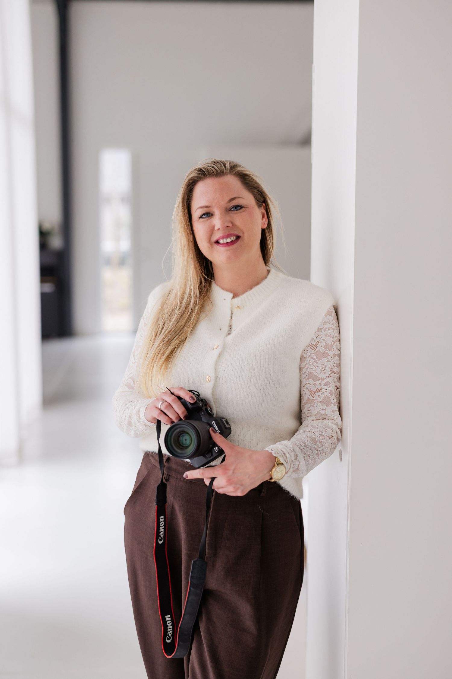 Smiling blonde woman photographer holding a Canon DSLR camera, wearing white lace top and brown trousers, leaning against wall.