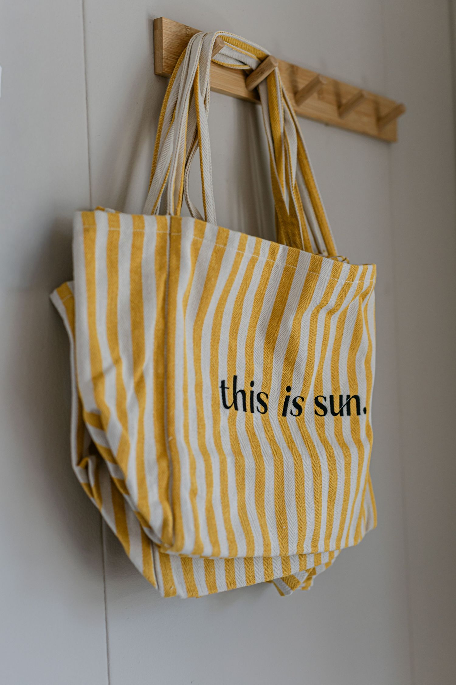 Yellow and white striped canvas tote bag with this is sun text hanging on wooden wall hook.