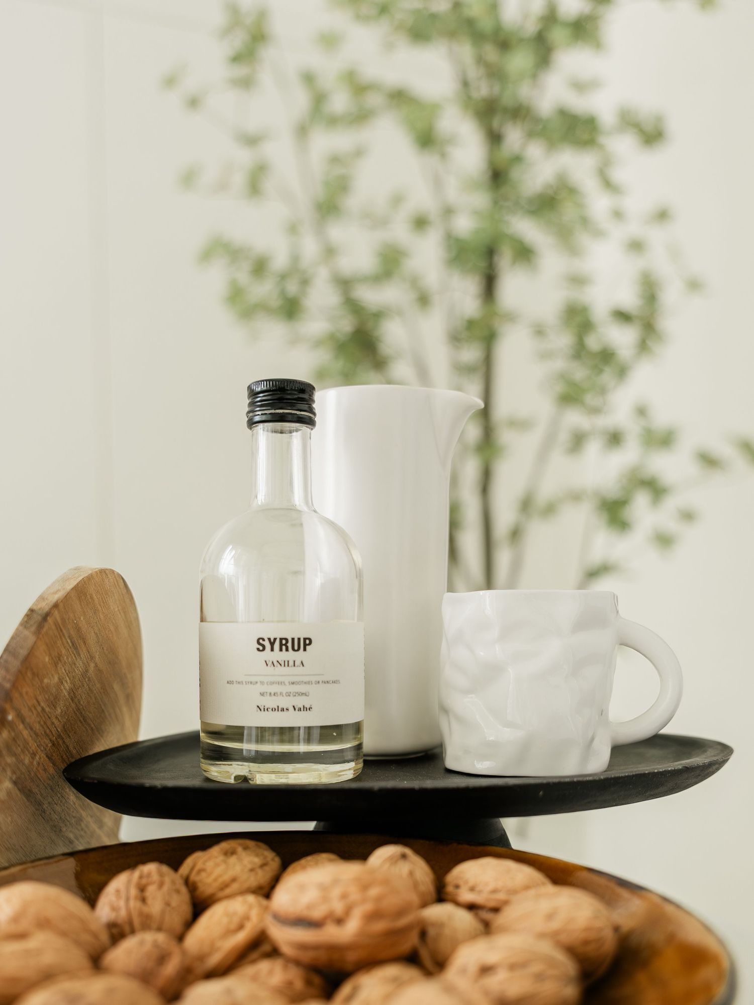 Minimalist flat lay with a glass syrup bottle, white ceramic mug, pitcher, and walnuts on a dark tray.