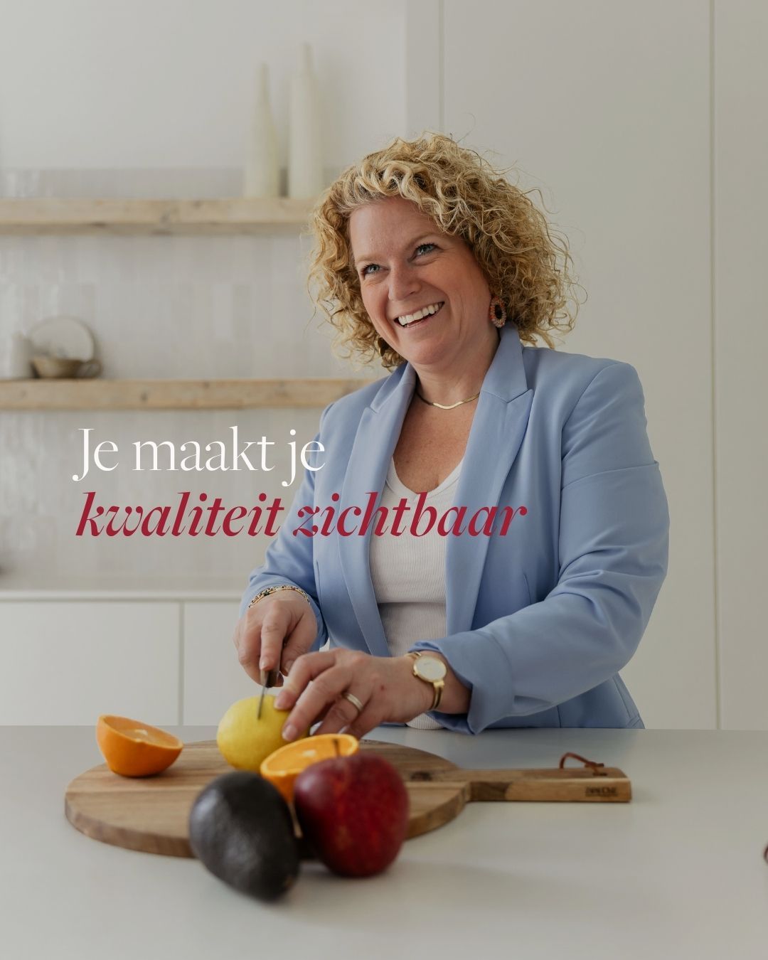 Curly-haired woman in blue blazer cutting fruit in modern kitchen with Dutch motivational text overlay.