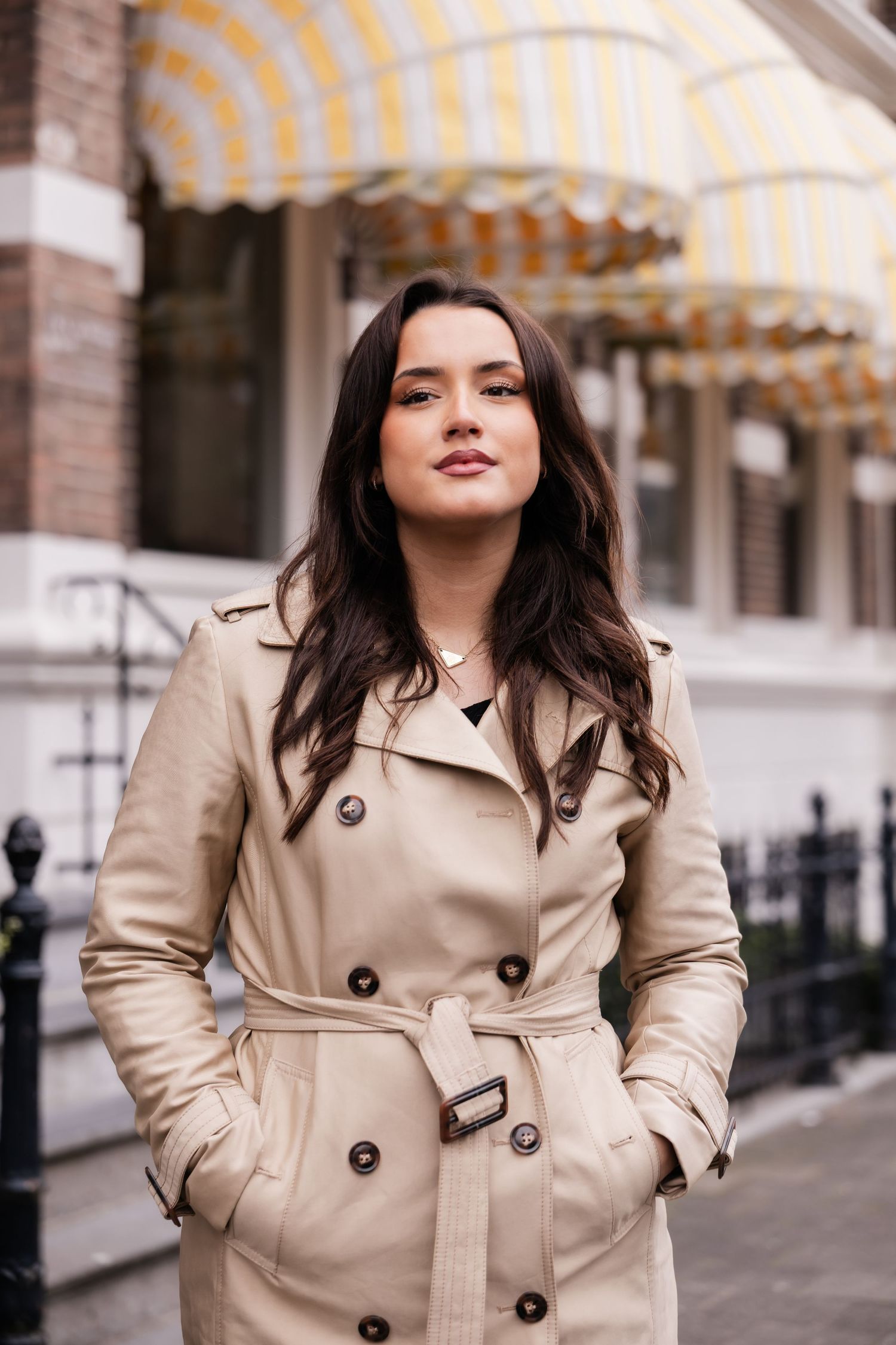 Confident brunette woman in belted beige trench coat on urban street.
