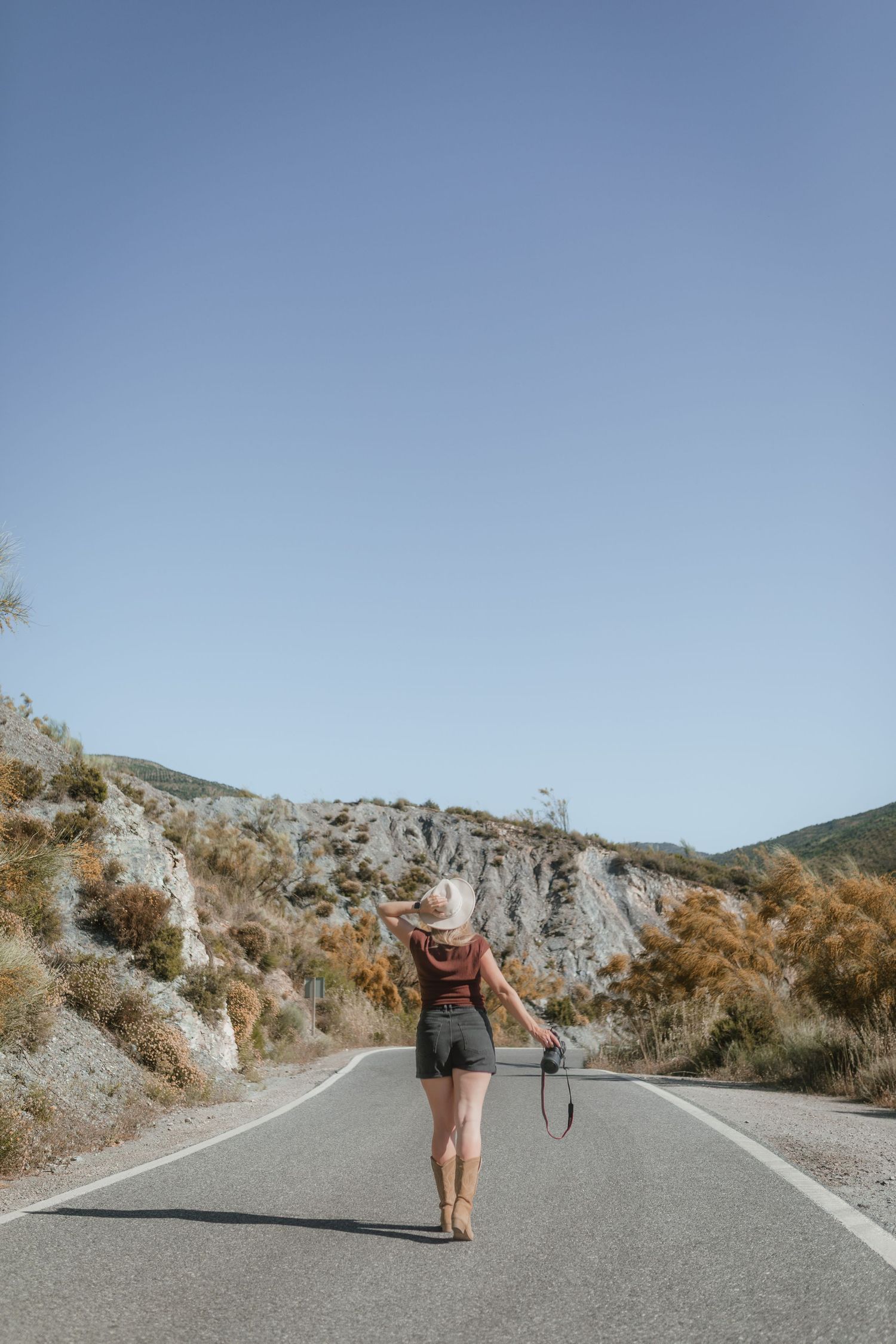 Vrouw in hoed en korte broek die over een schilderachtige bergweg loopt onder een heldere blauwe lucht.