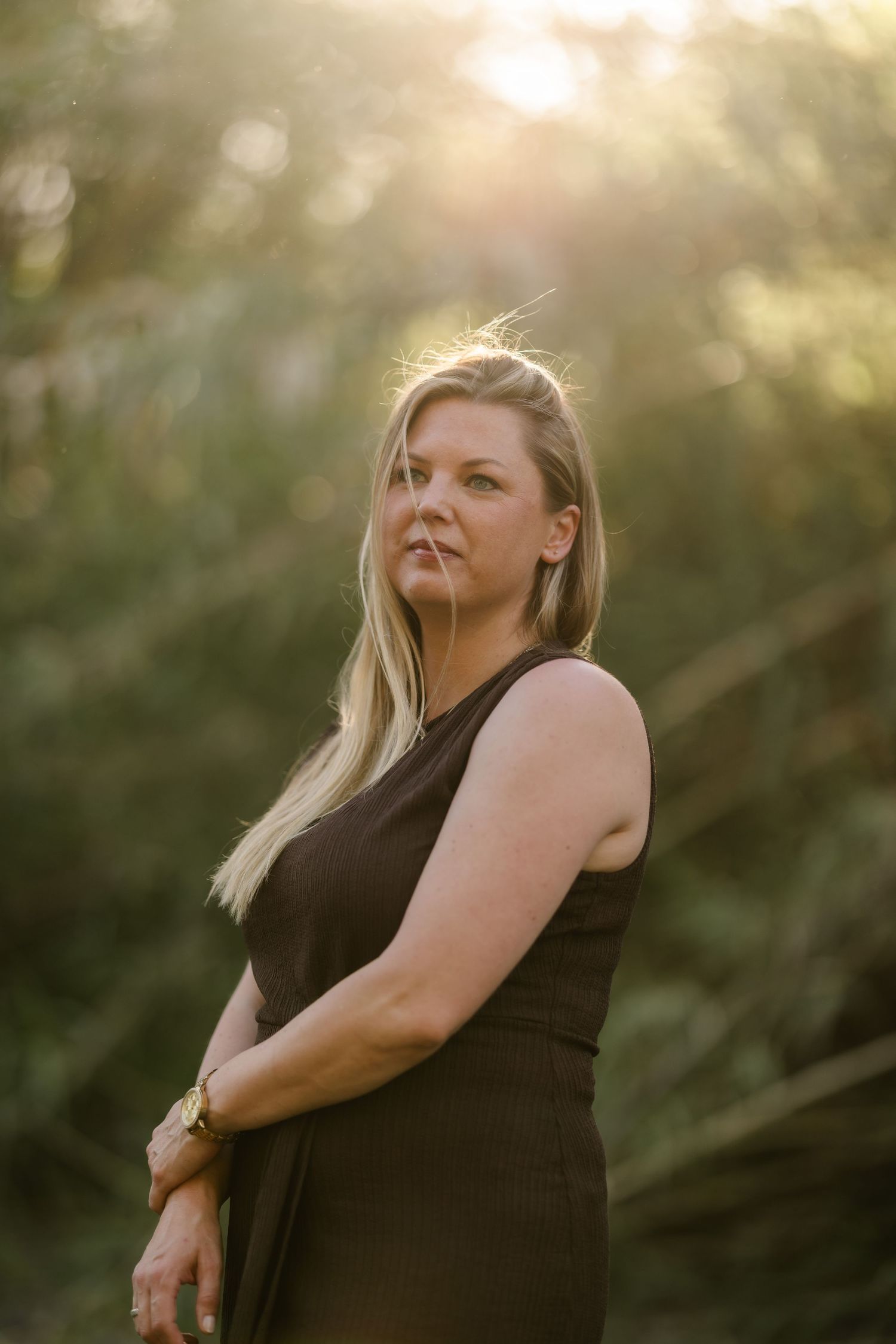 Blonde woman in black sleeveless dress poses outdoors at golden hour with sunlit green trees in background.