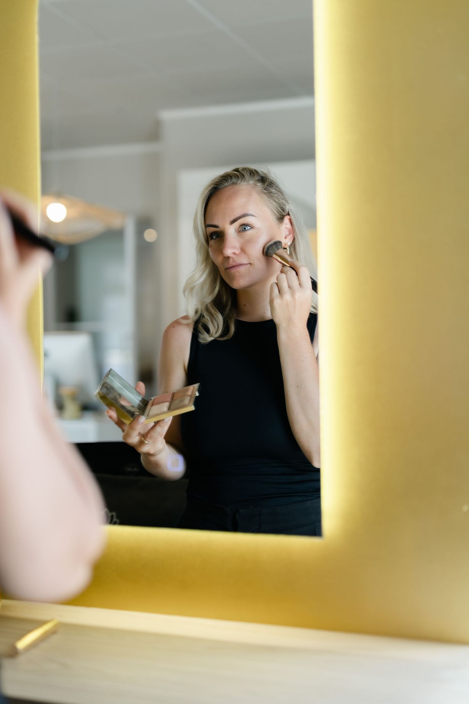 Blonde woman applying bronzer with brush while looking in yellow-framed illuminated mirror.