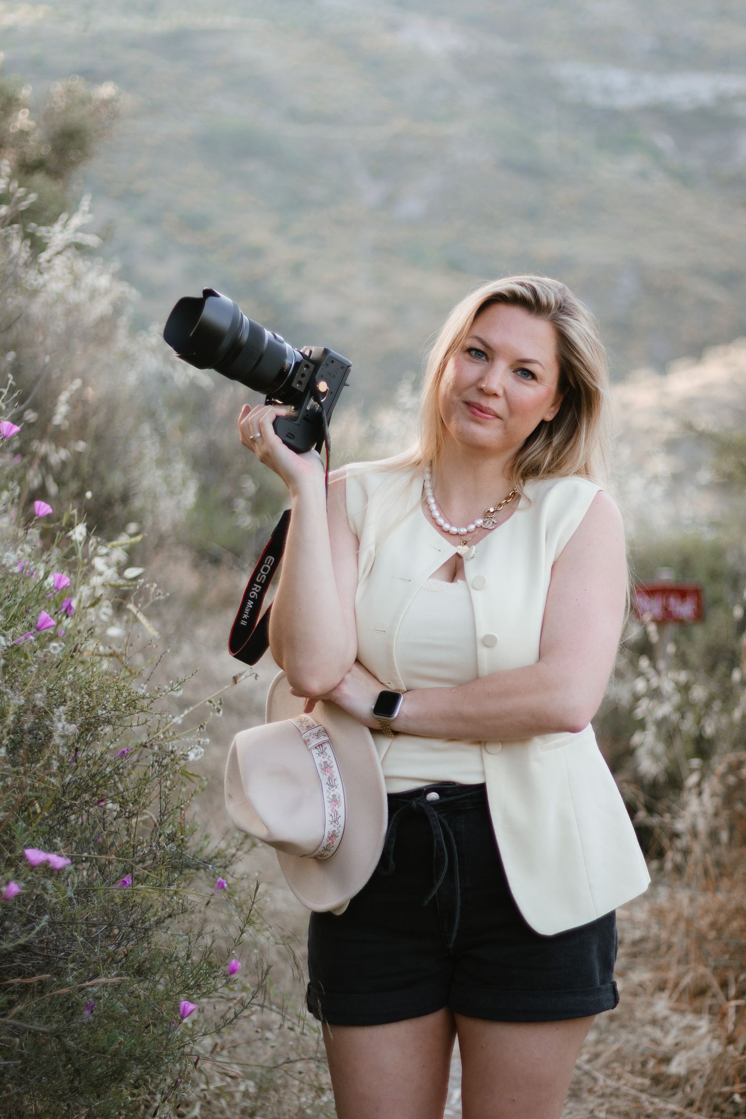 Blonde woman holding a DSLR camera and hat outdoors in a scenic mountain landscape with wildflowers.