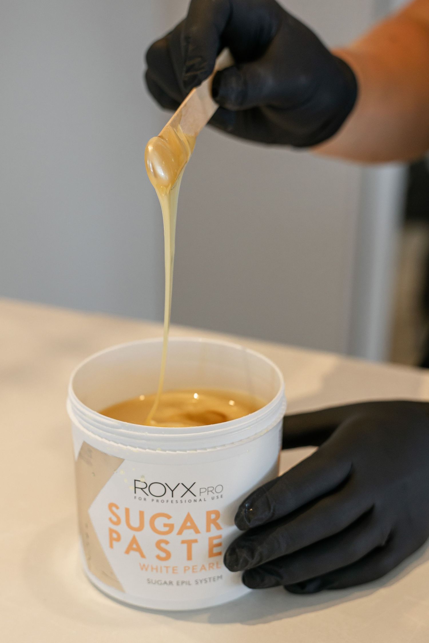 Gloved hands using spatula to scoop ROYX Pro Sugar Paste White Pearl wax from container.