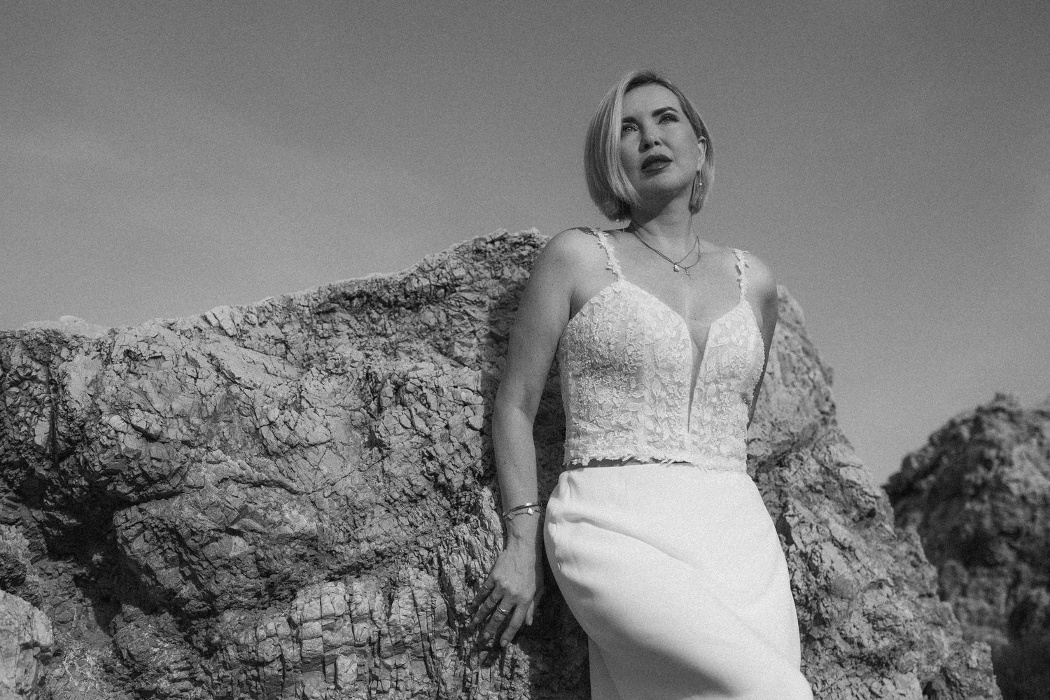 Vrouw in witte kanten top en rok poseert tegen rotsachtige kliffen in elegante zwart-wit buitenfoto.