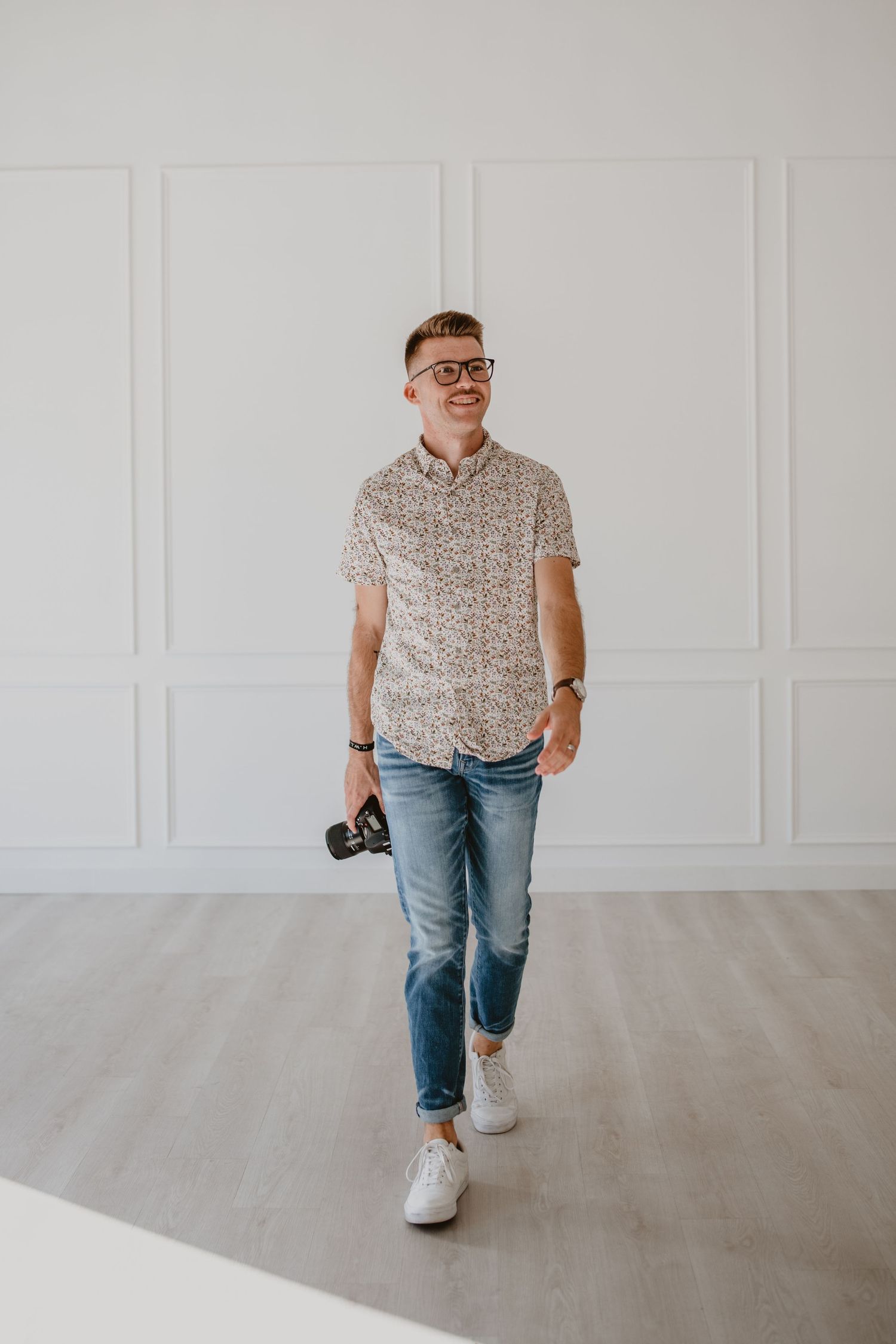 A photographer in casual attire walks while holding a camera in a bright white studio with hardwood floors.