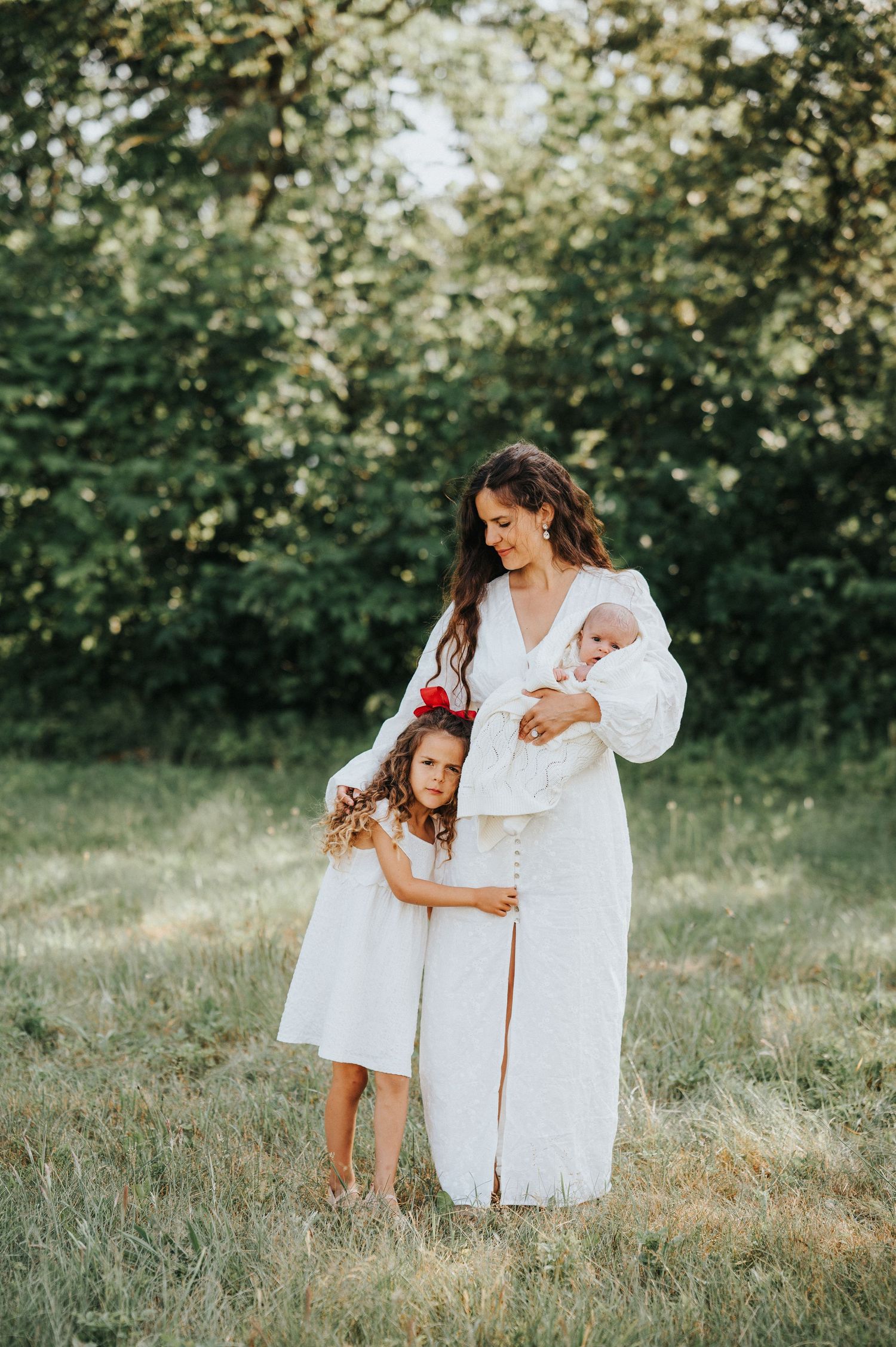 Mother in white dress holds baby while young daughter in white dress stands beside her outdoors.