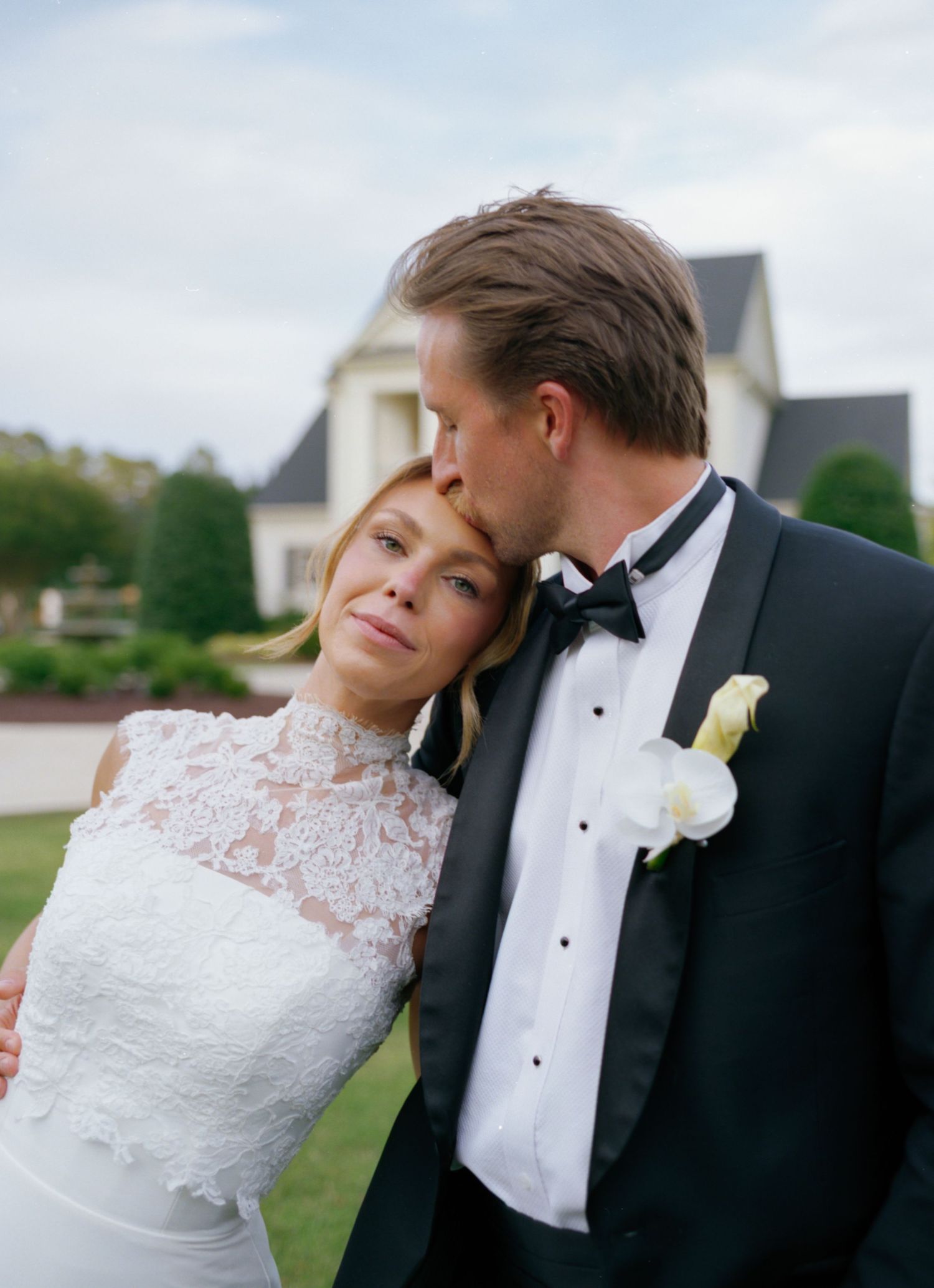 A bride in white lace gown leans on groom in black tuxedo with white boutonniere as he kisses her forehead outdoors.