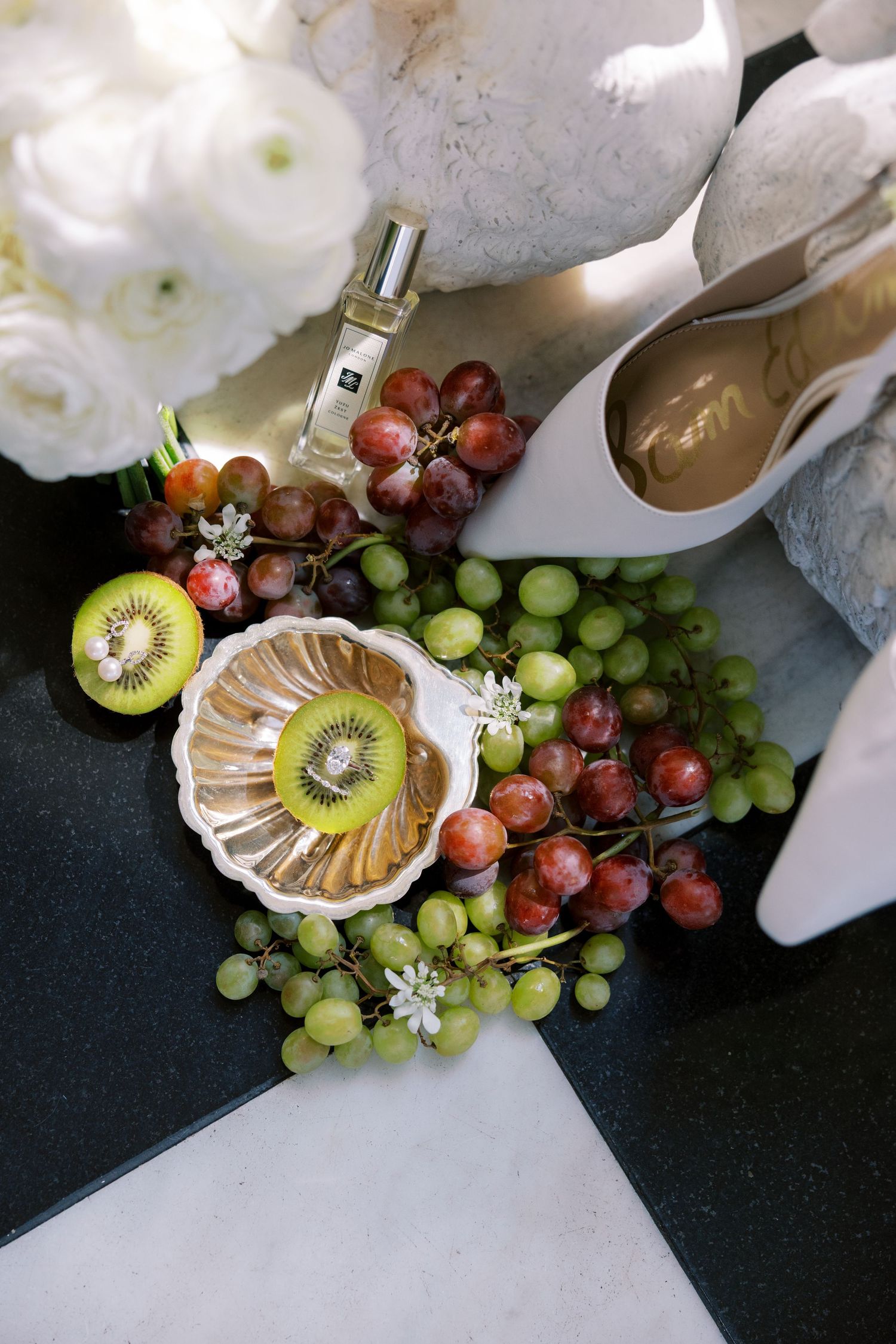 Elegant bridal flat lay with white heels, Jo Malone perfume, kiwi, red and green grapes, and white ranunculus bouquet.