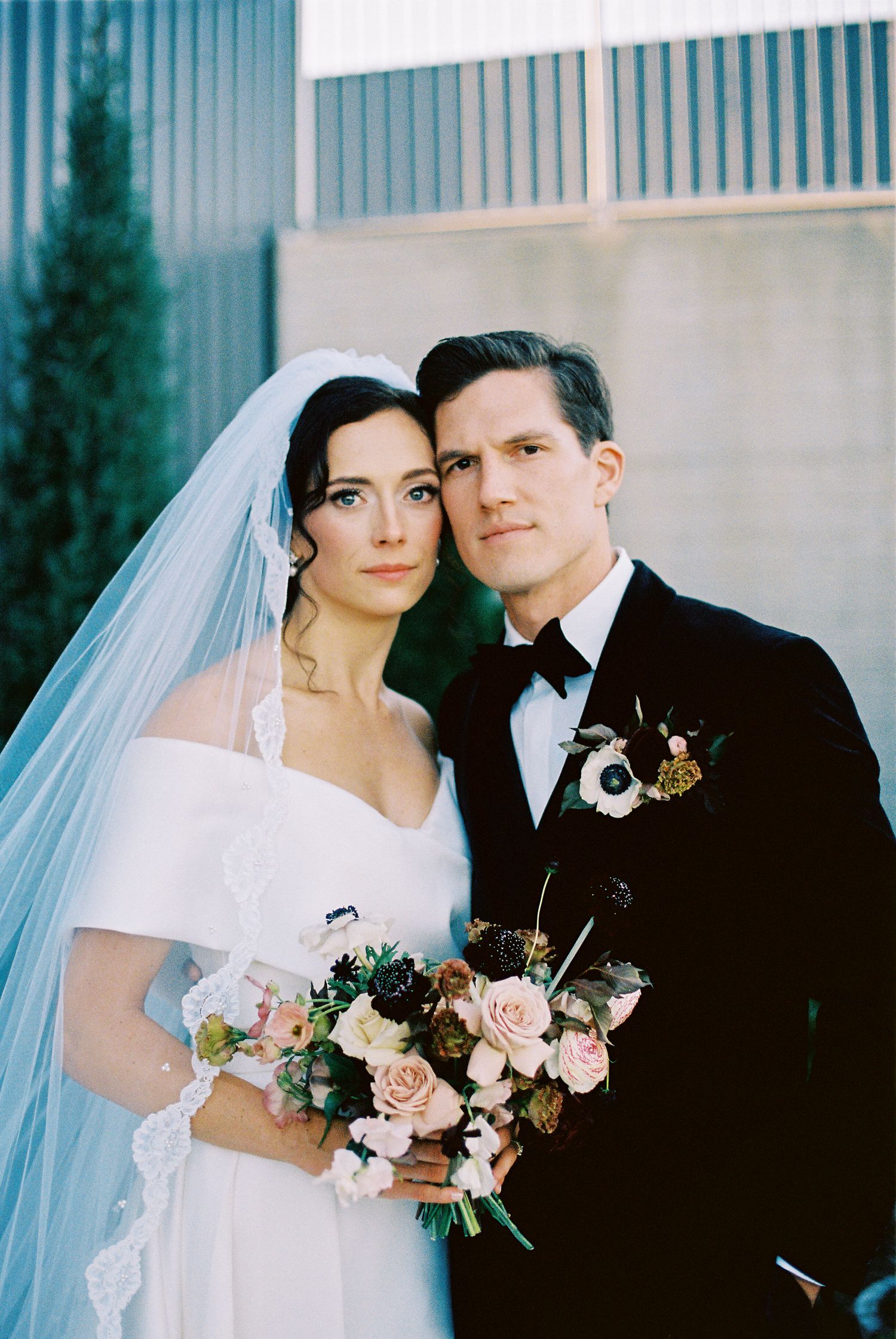 A bride and groom pose together at their wedding, with the bride holding a pink and white floral bouquet.